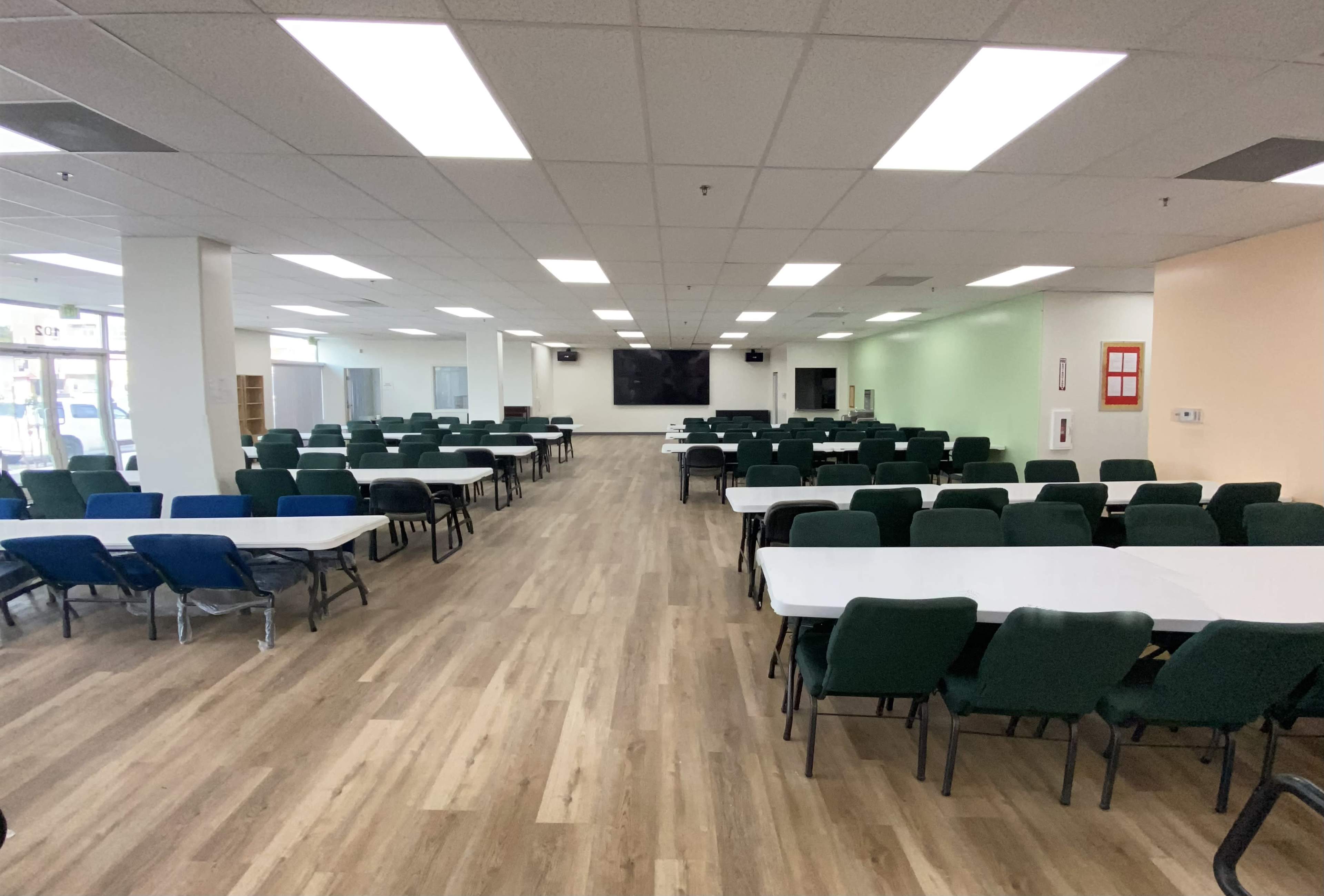 A large room with multiple rows of tables and chairs arranged for dining or meetings, featuring bright overhead lighting and wooden flooring.