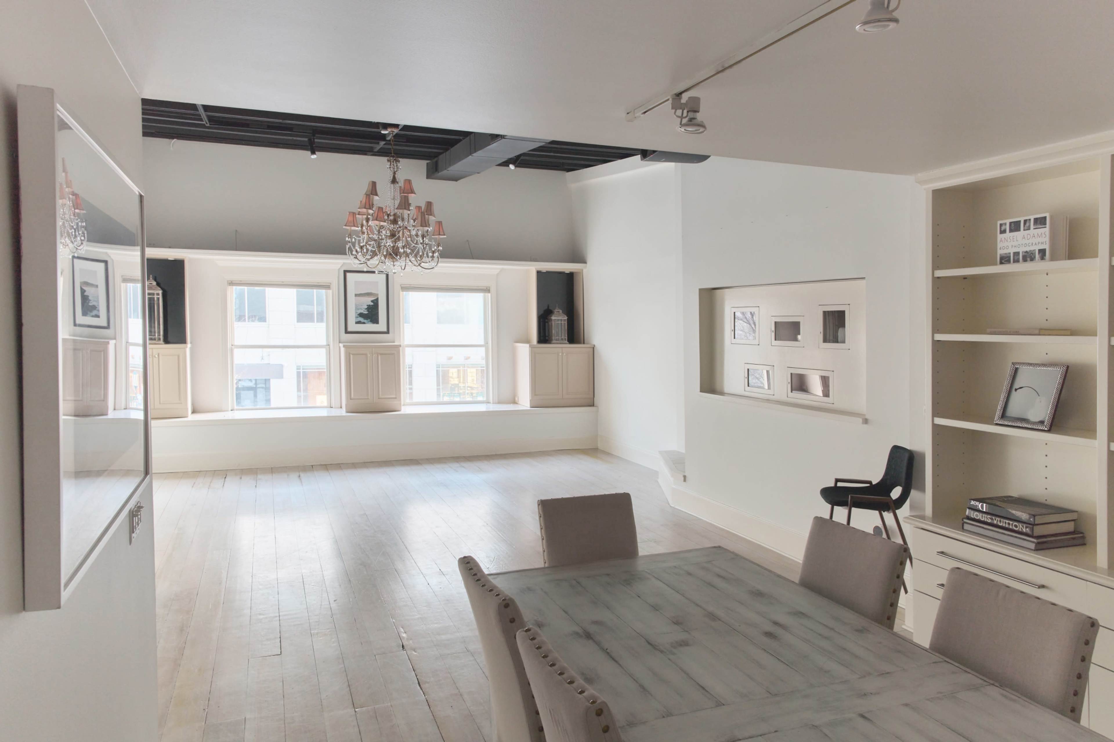 The image shows a spacious, modern room with white walls, wooden flooring, a dining table, and a chandelier, featuring large windows with built-in seating.