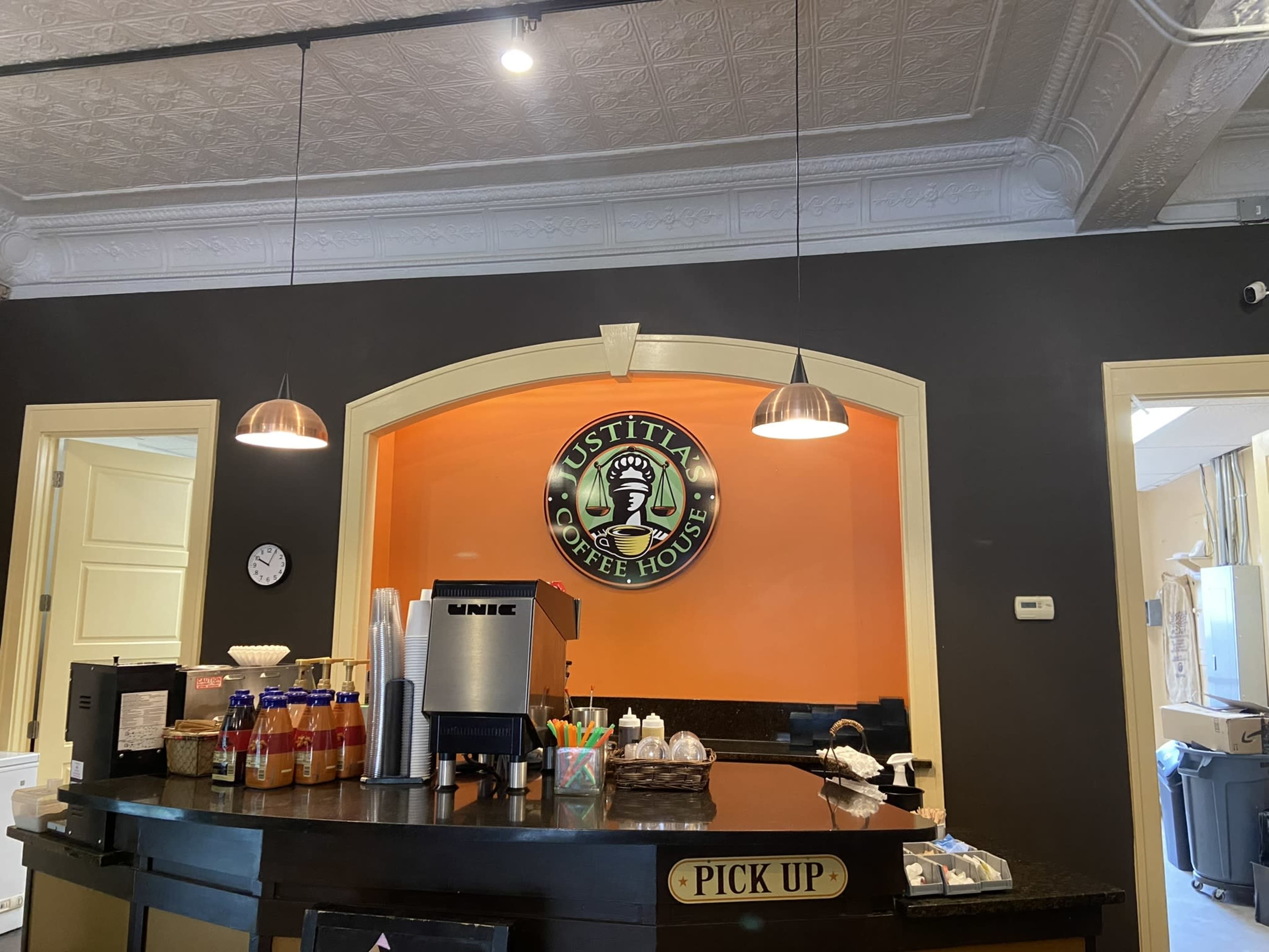 The image shows the interior of a coffee house with a service counter, a circular logo on the wall, and pendant lights overhead.