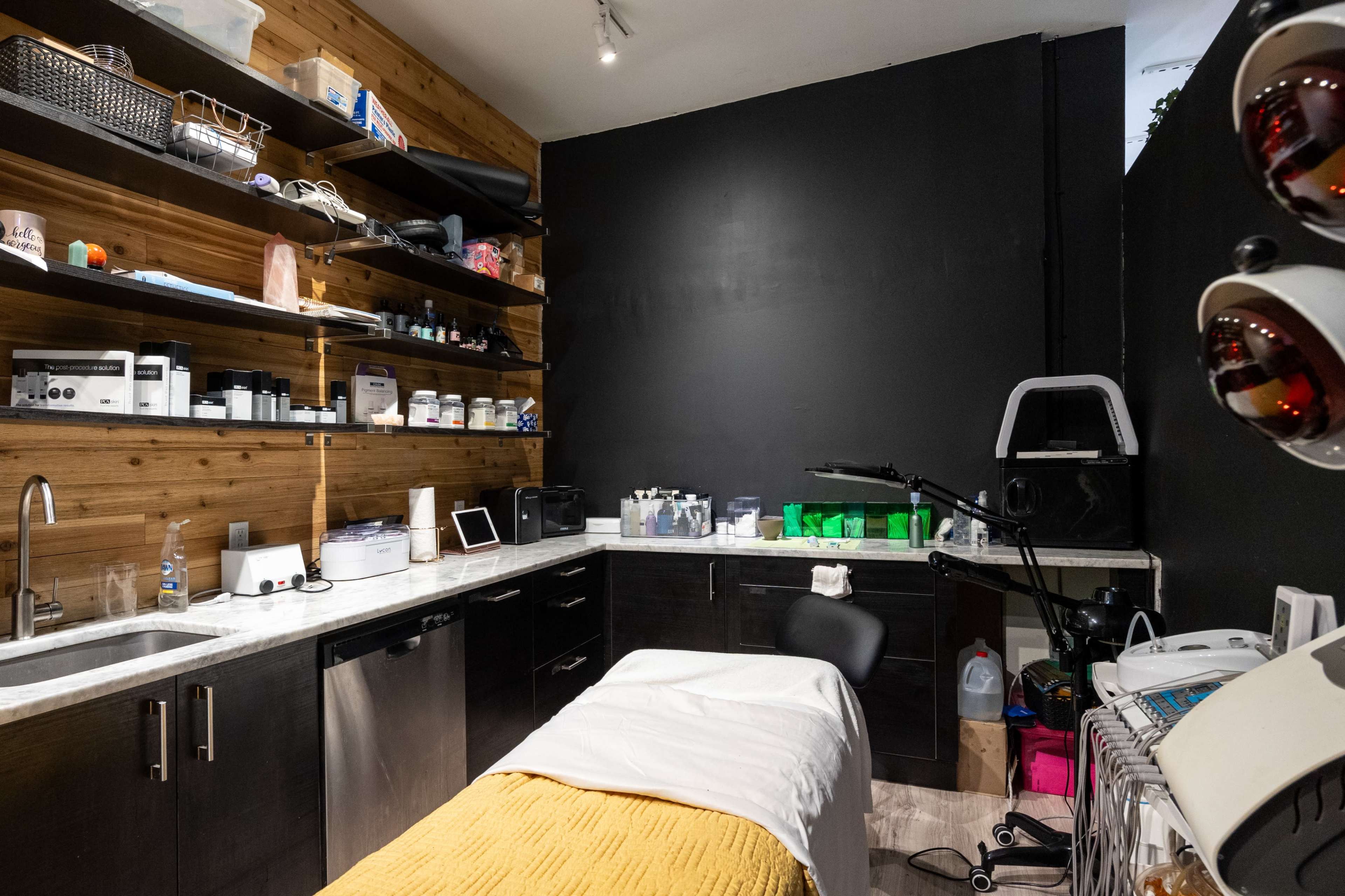 A compact treatment room with a dark wall, shelves holding various beauty products, a treatment bed covered with a white sheet, and a sink with cabinets beneath.