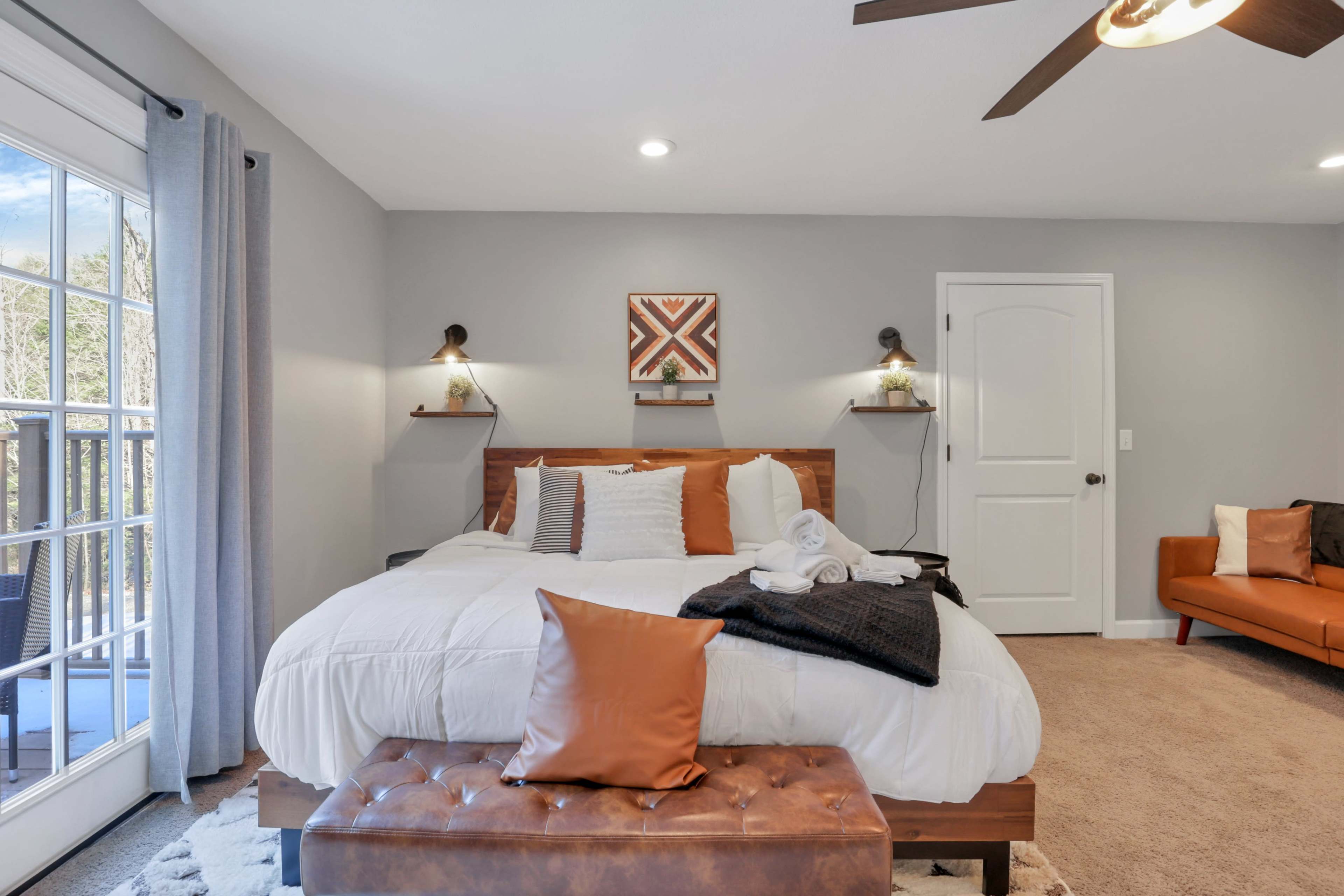 A well-decorated bedroom with a king-sized bed, a brown leather bench at the foot, and a small orange couch in the corner, along with large windows that let in natural light.