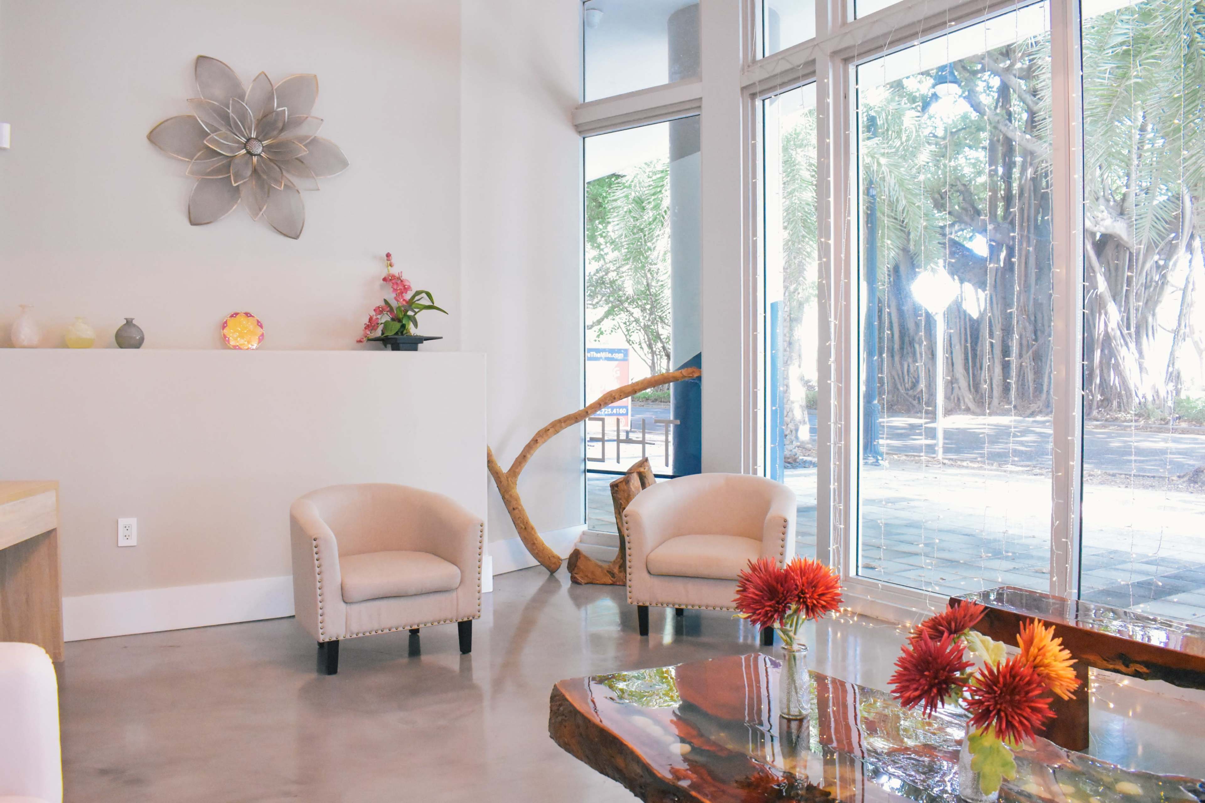 A modern interior space with two beige armchairs, a wooden coffee table, and large windows displaying greenery outside.