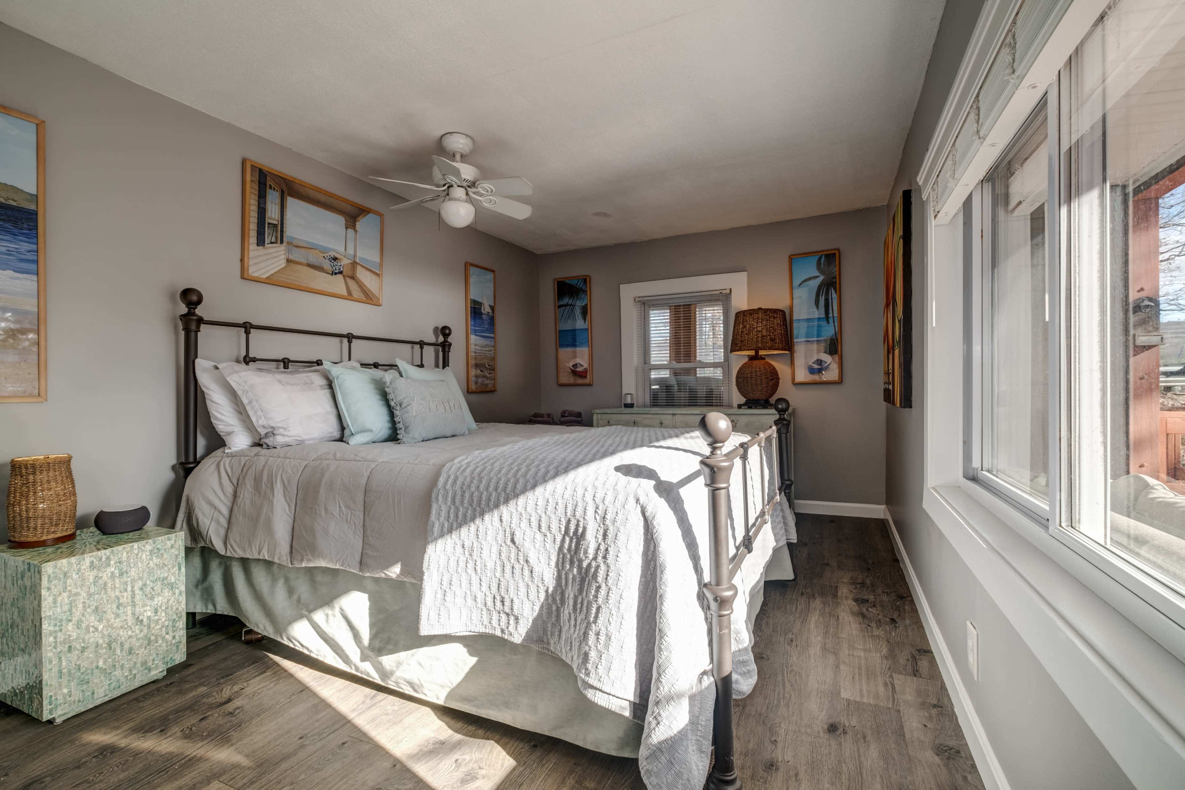 A well-lit bedroom features a metal bed frame with white bedding, gray walls adorned with artwork, and large windows offering natural light.