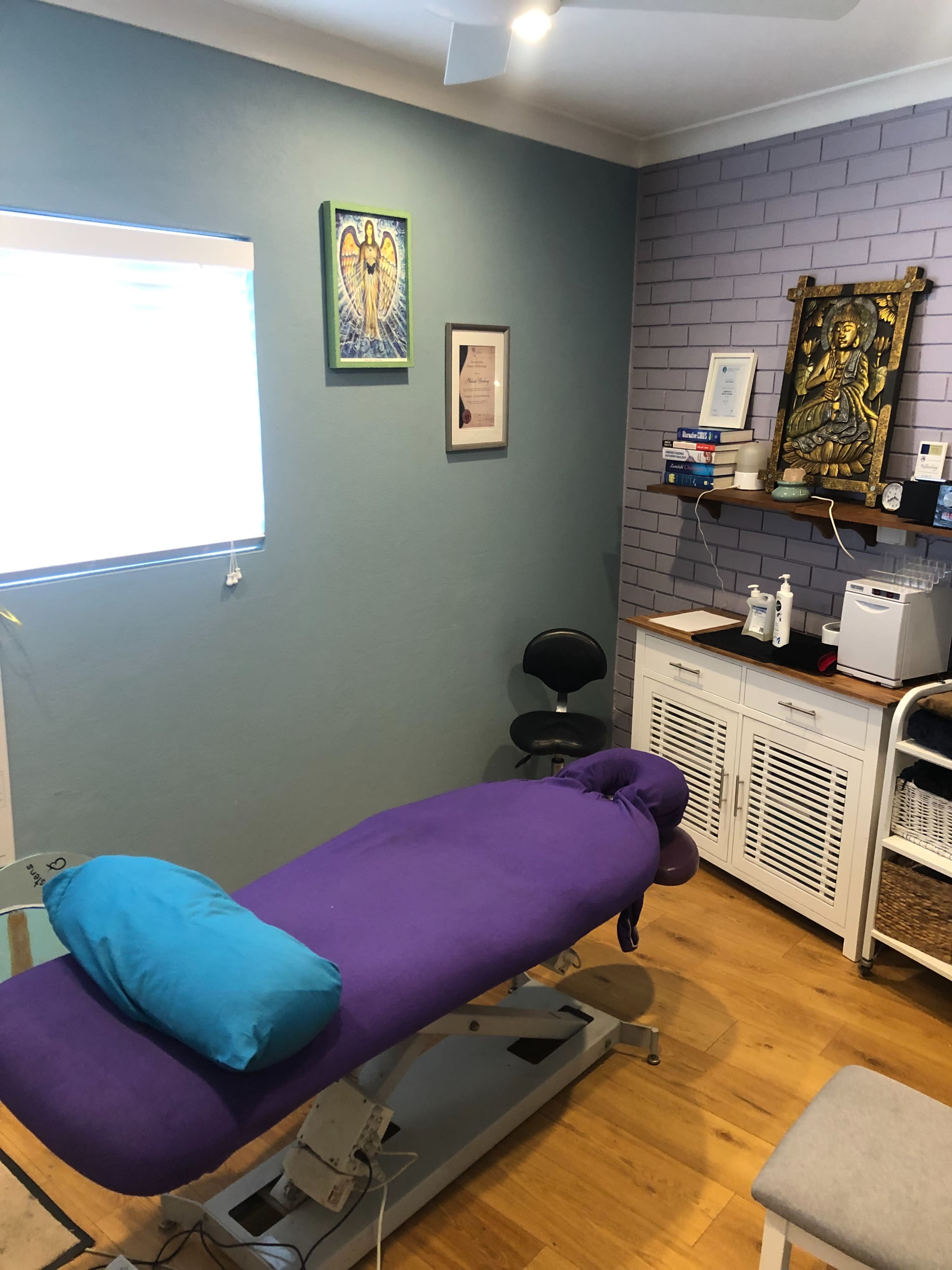 The image shows a therapy room featuring a purple massage table, a chair, a shelf with various supplies, and artwork on the walls.