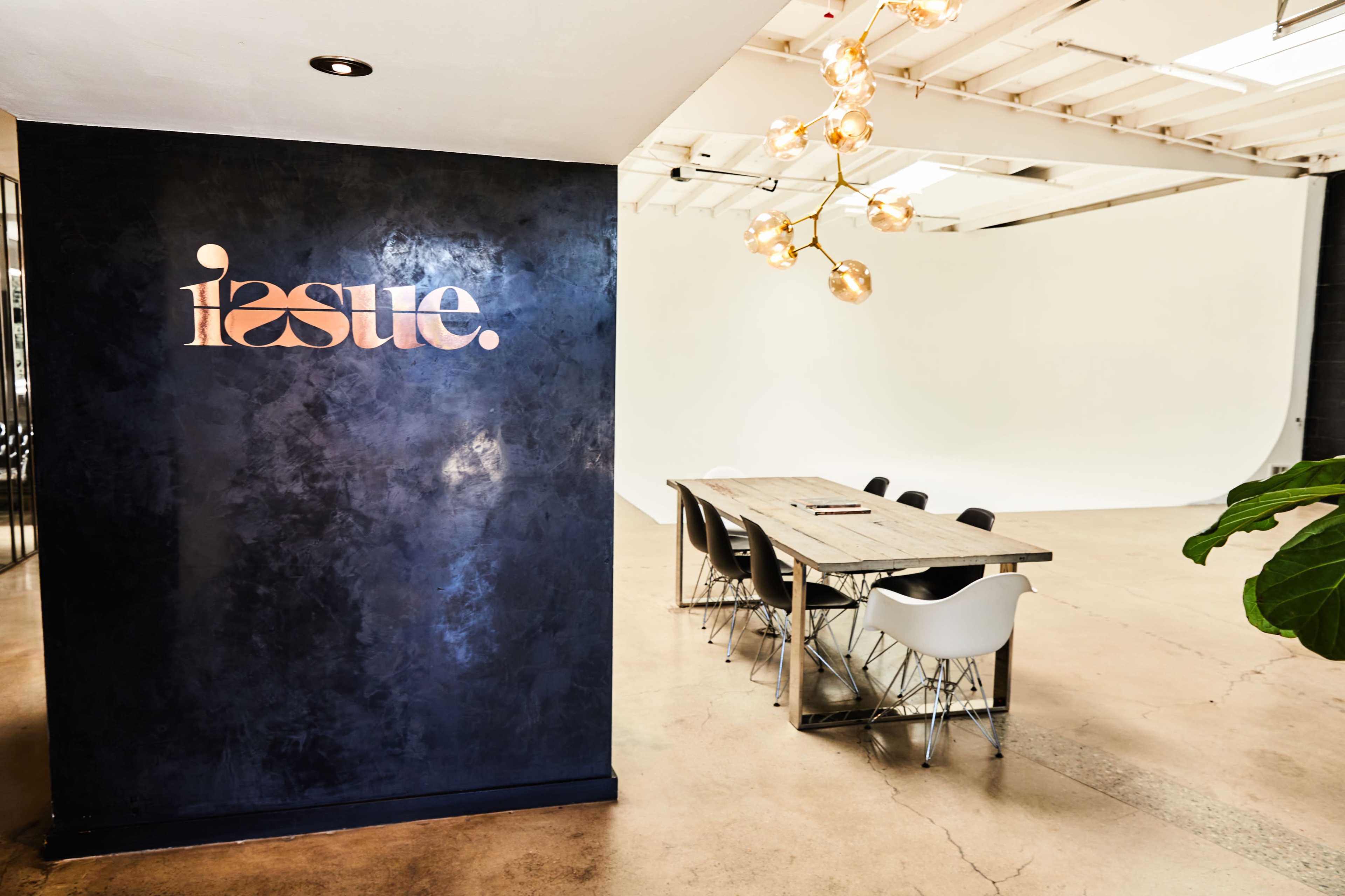A modern office space with a black wall featuring the word "issue" and a long wooden table surrounded by white chairs.