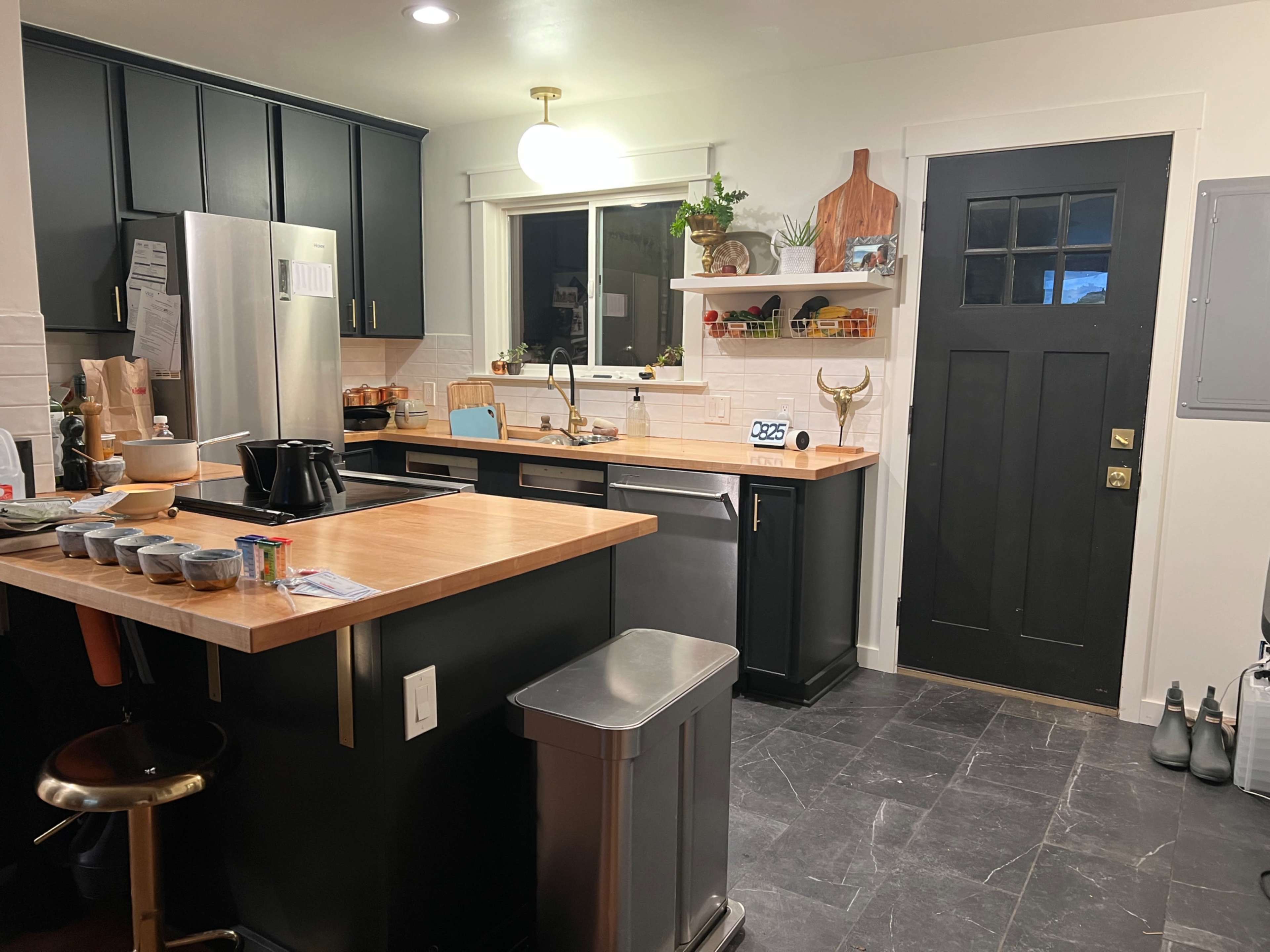 The image shows a modern kitchen with black cabinets, a wooden countertop, stainless steel appliances, and an adjacent door leading outside.