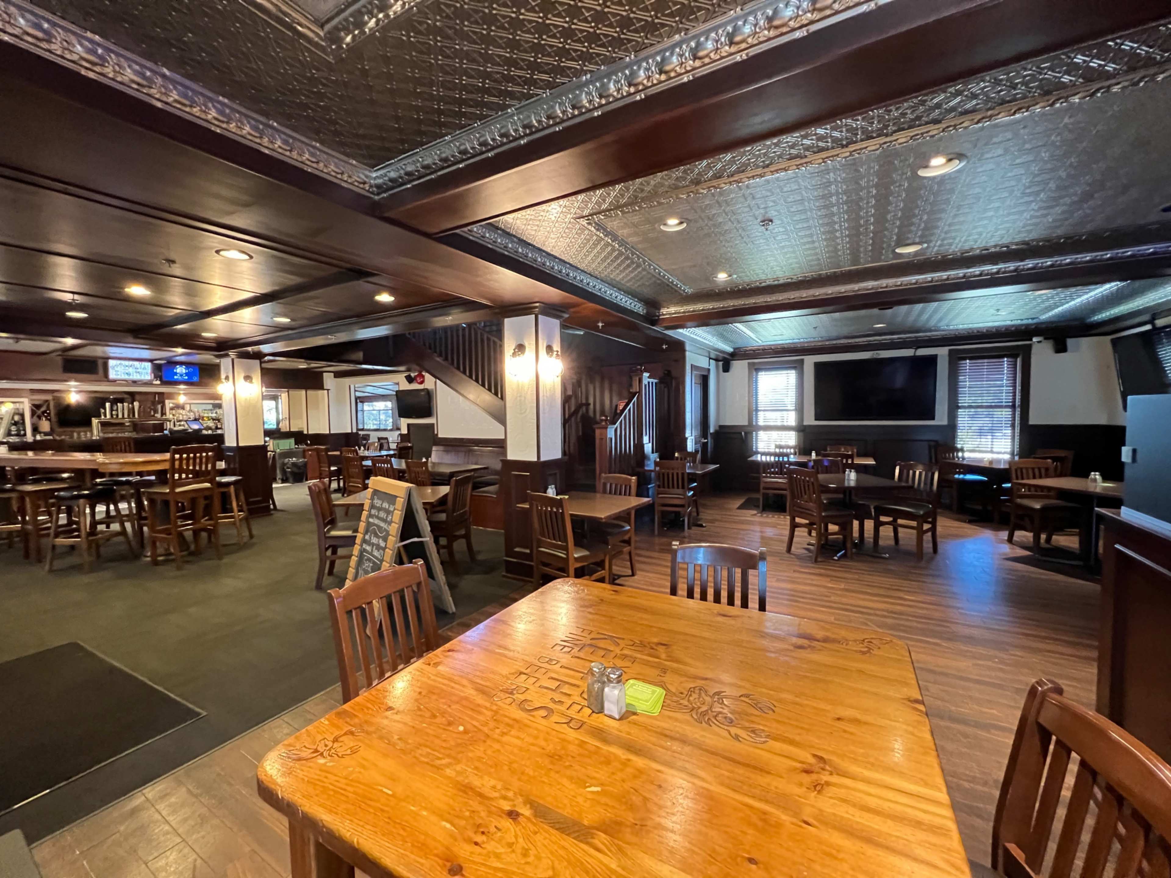 The interior of a restaurant with wooden furniture, a bar area, and a mix of empty tables and chairs.