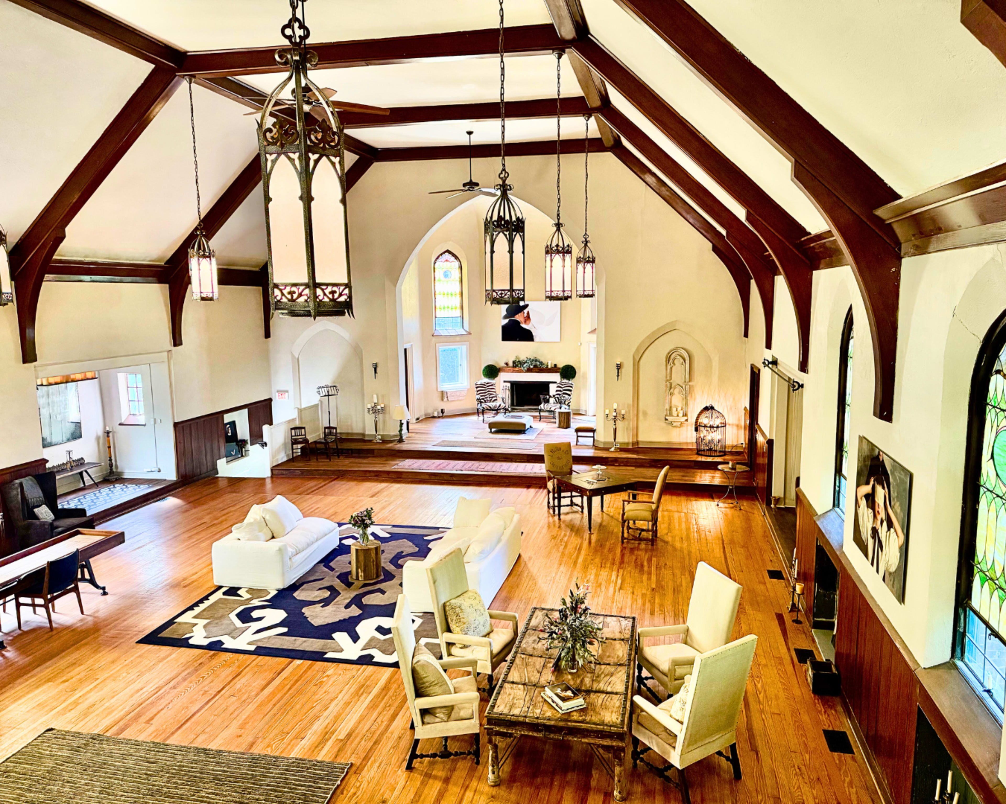 The image shows a spacious, well-lit living area with high wooden beams, a large rug, and a mix of modern and traditional furnishings in a church-turned-home setting.