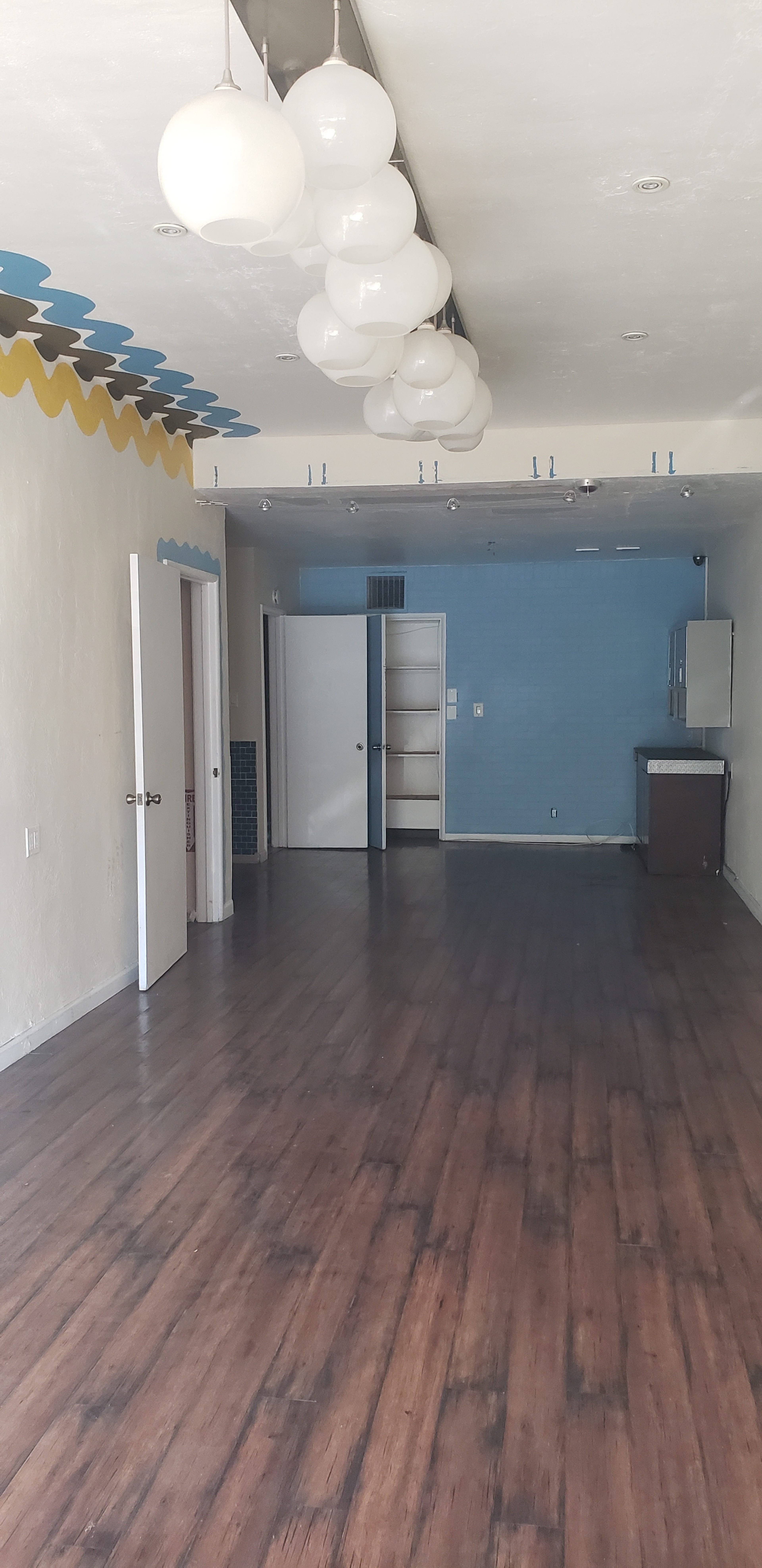 The image shows an empty room with wooden floors, white walls, and large pendant lights hanging from the ceiling, leading to an open area with multiple doors.