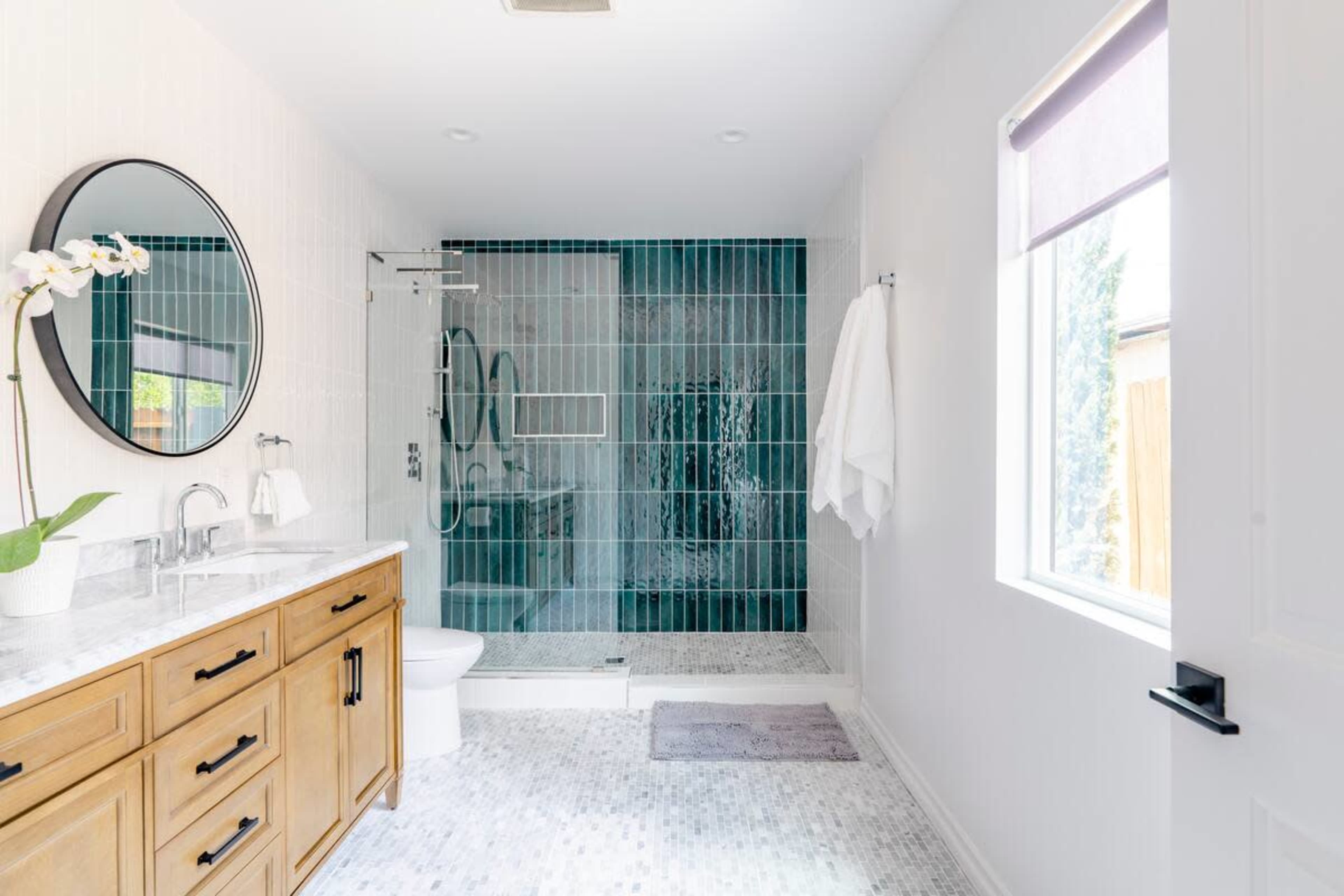 A modern bathroom featuring a glass-enclosed shower with teal tiles, a wooden vanity with a marble countertop, and a circular mirror.