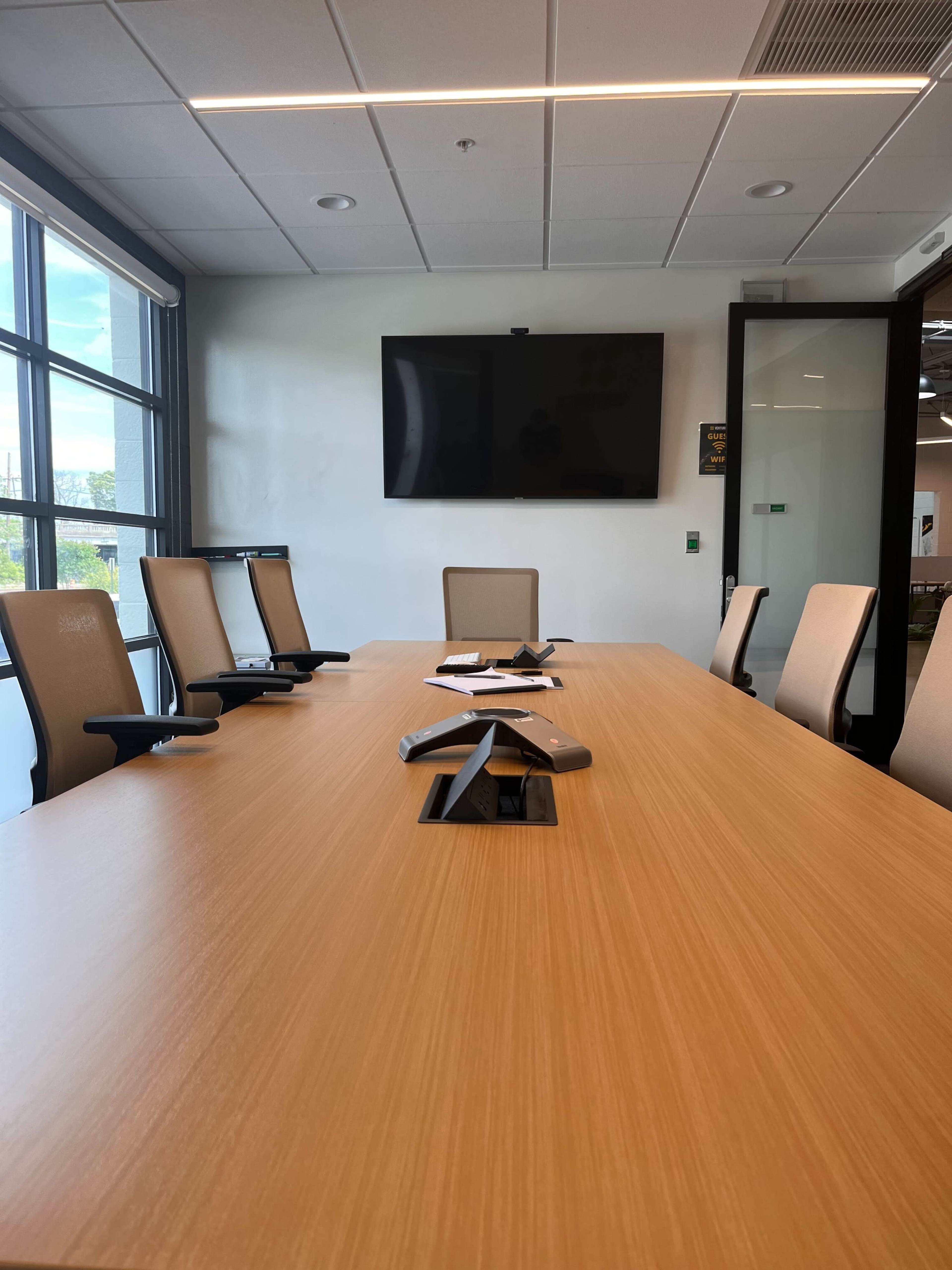 A modern conference room features a large wooden table with several chairs and a wall-mounted TV.