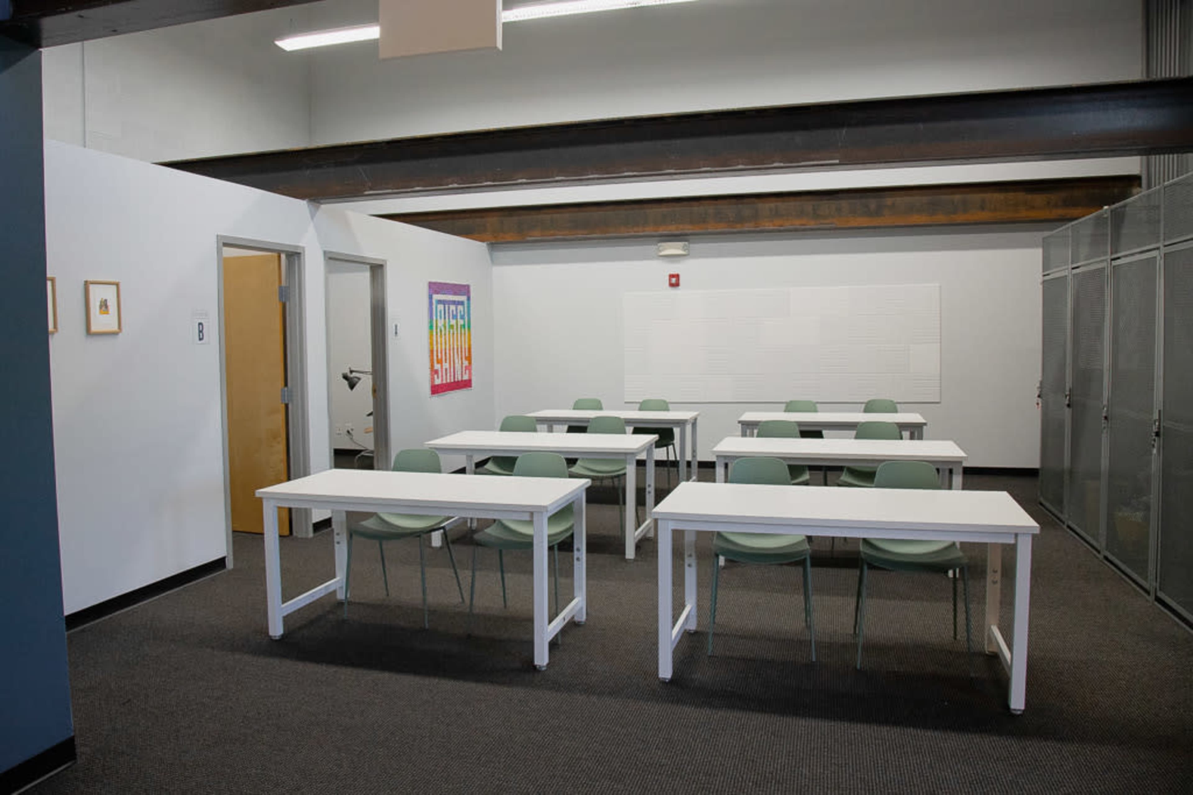 A classroom with several white tables and green chairs arranged in a minimalist layout against a gray wall.