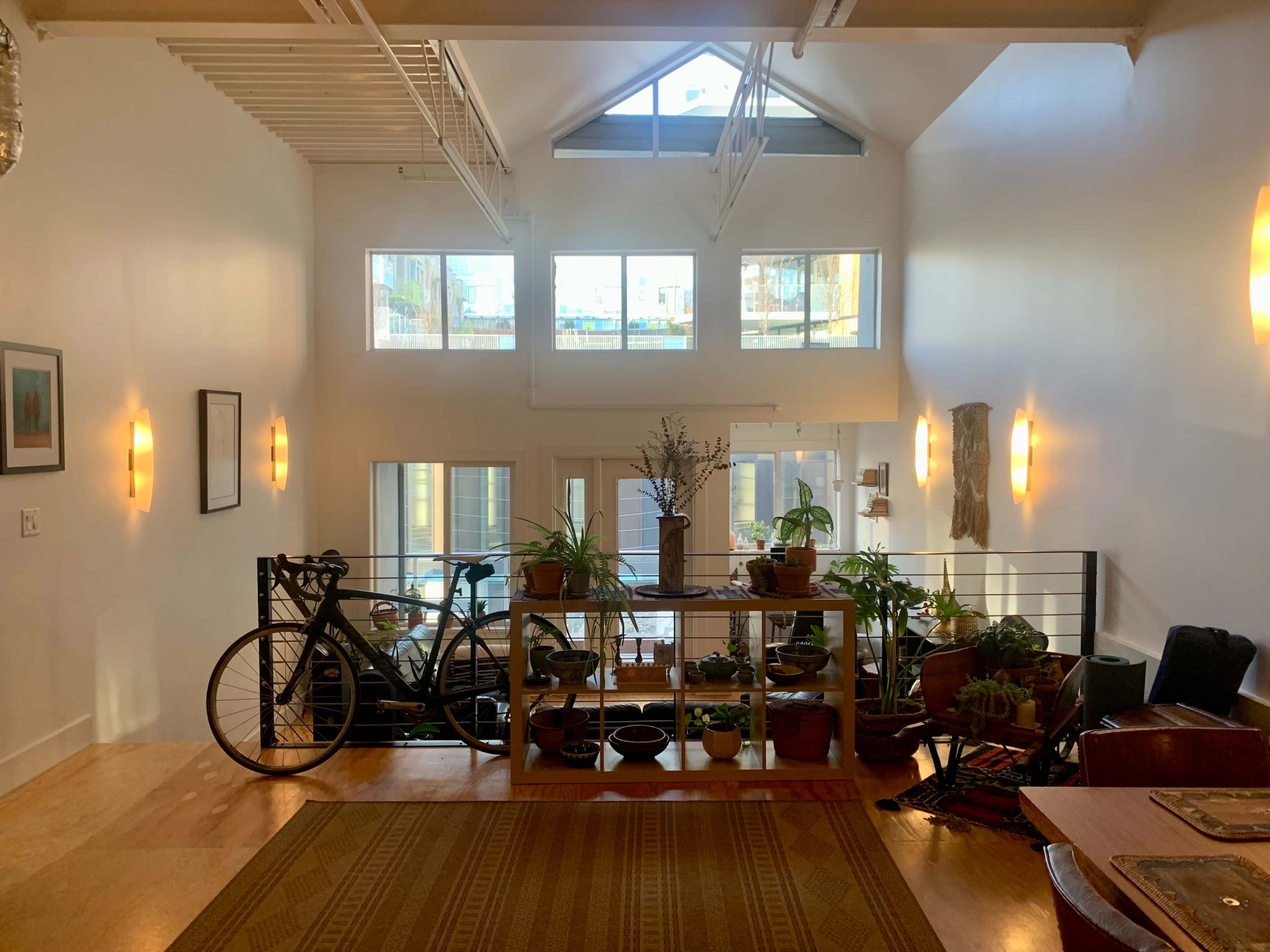 A spacious entryway with large windows, a bicycle leaning against a wooden shelf filled with plants, and a view of an open living area beyond.