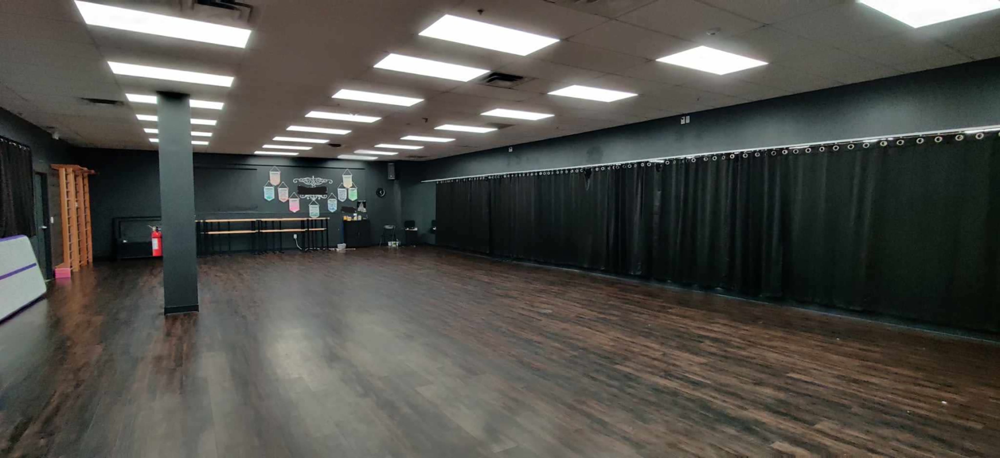 A spacious indoor room with dark wooden flooring, black curtains along one wall, and bright ceiling lights illuminating the area.
