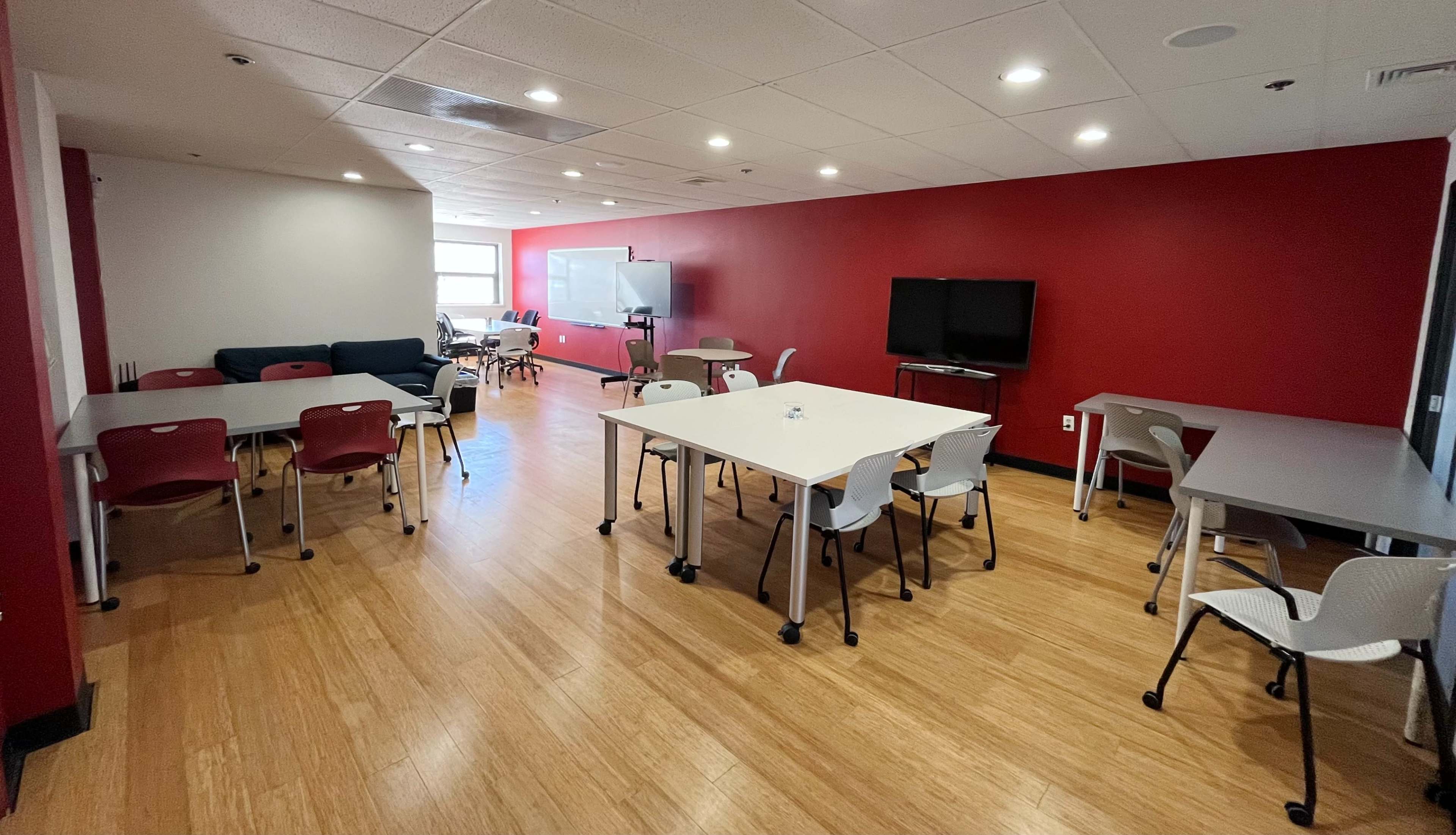 The image shows a well-lit room with multiple tables and chairs, a sofa in one corner, and two screens on opposite walls, set against a red-painted backdrop.
