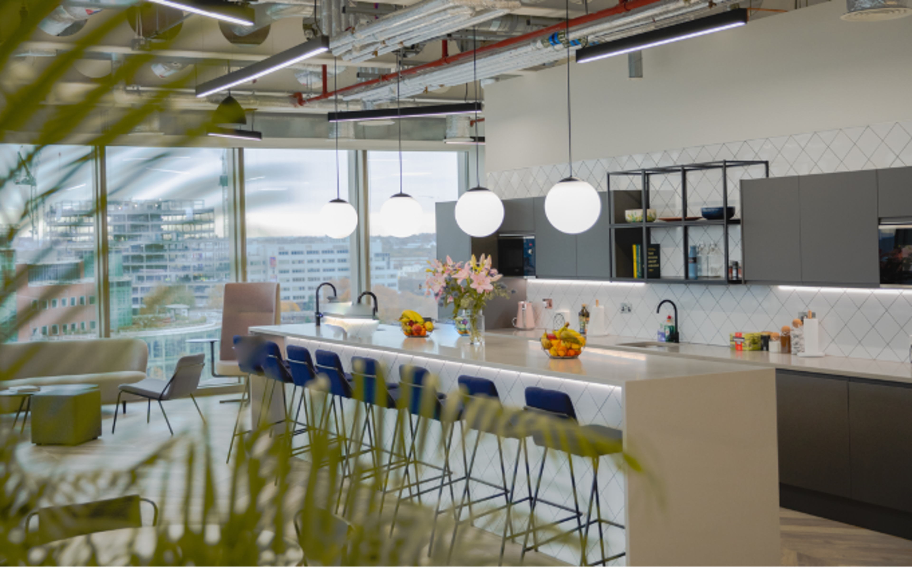 A modern kitchen area features a long countertop with round light fixtures overhead and views of the city through large windows.