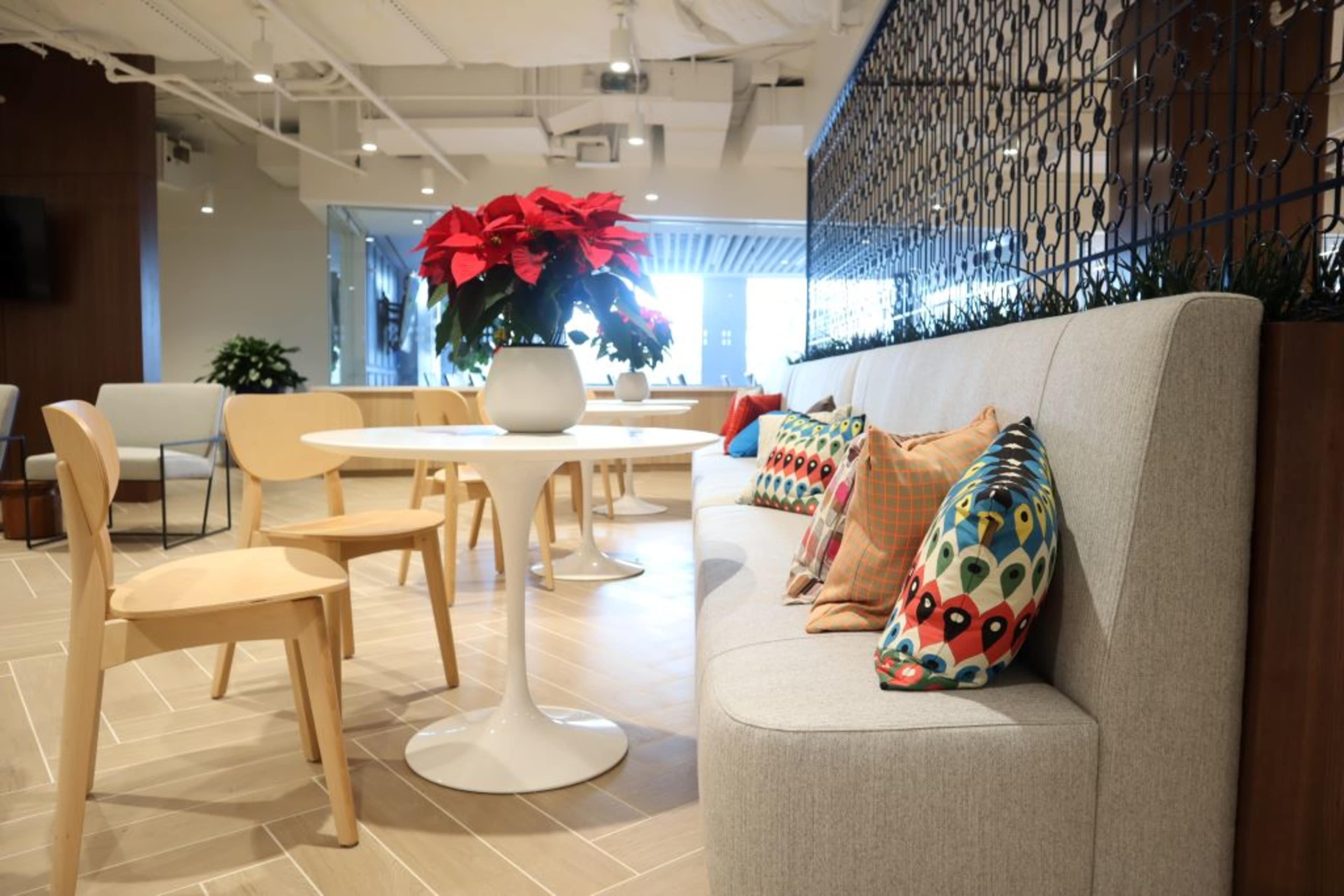 A modern seating area features a gray upholstered bench with colorful pillows, a round white table, wooden chairs, and a poinsettia plant in a white vase.
