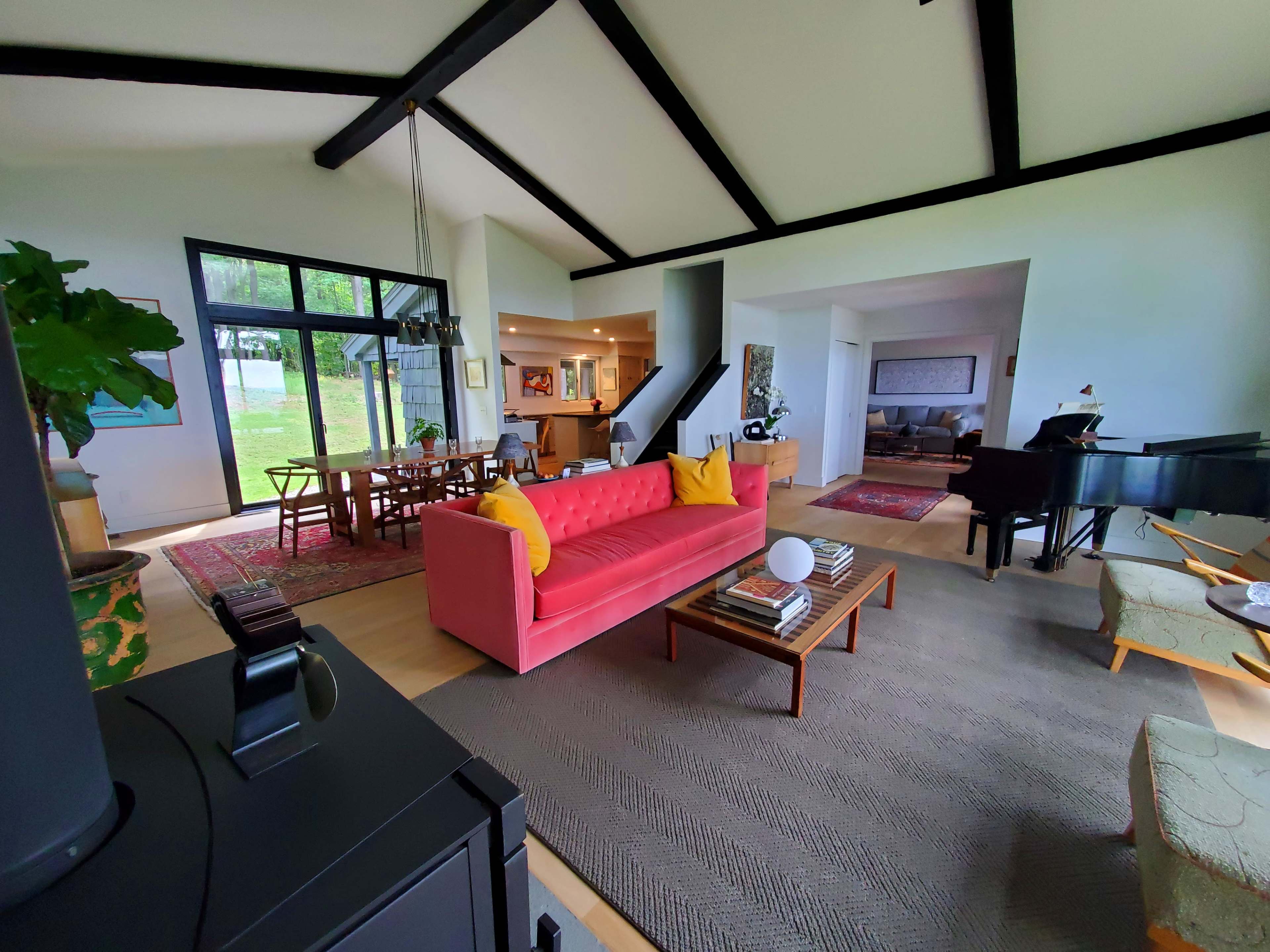 The image shows a spacious living area featuring a red sofa, a dining table, and a piano, with large windows that provide a view of the outdoors.