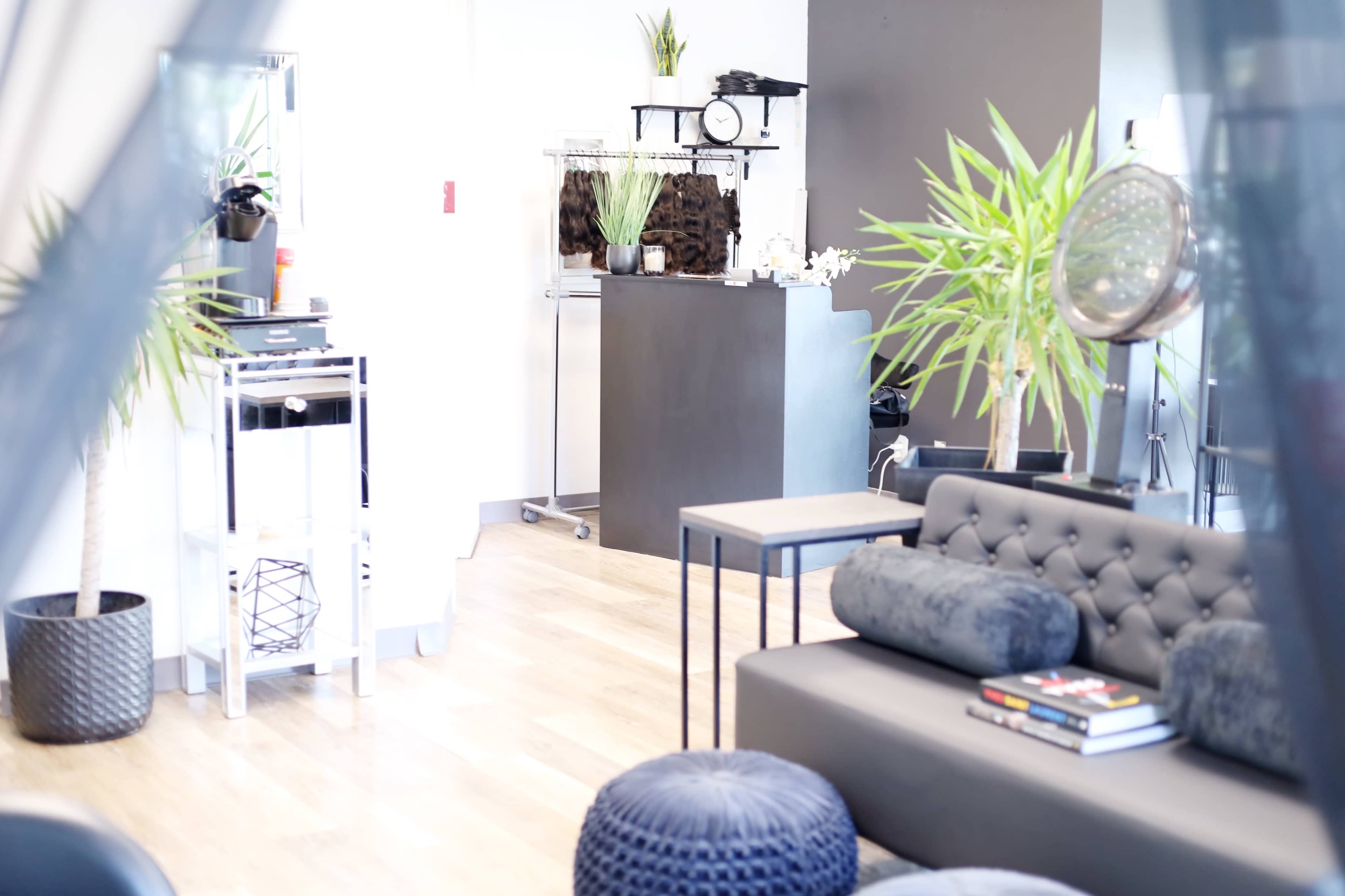 The image shows a modern interior space featuring a seating area with a dark sofa and round poufs, a small table, and a clothing rack in the background against a light-colored wall.
