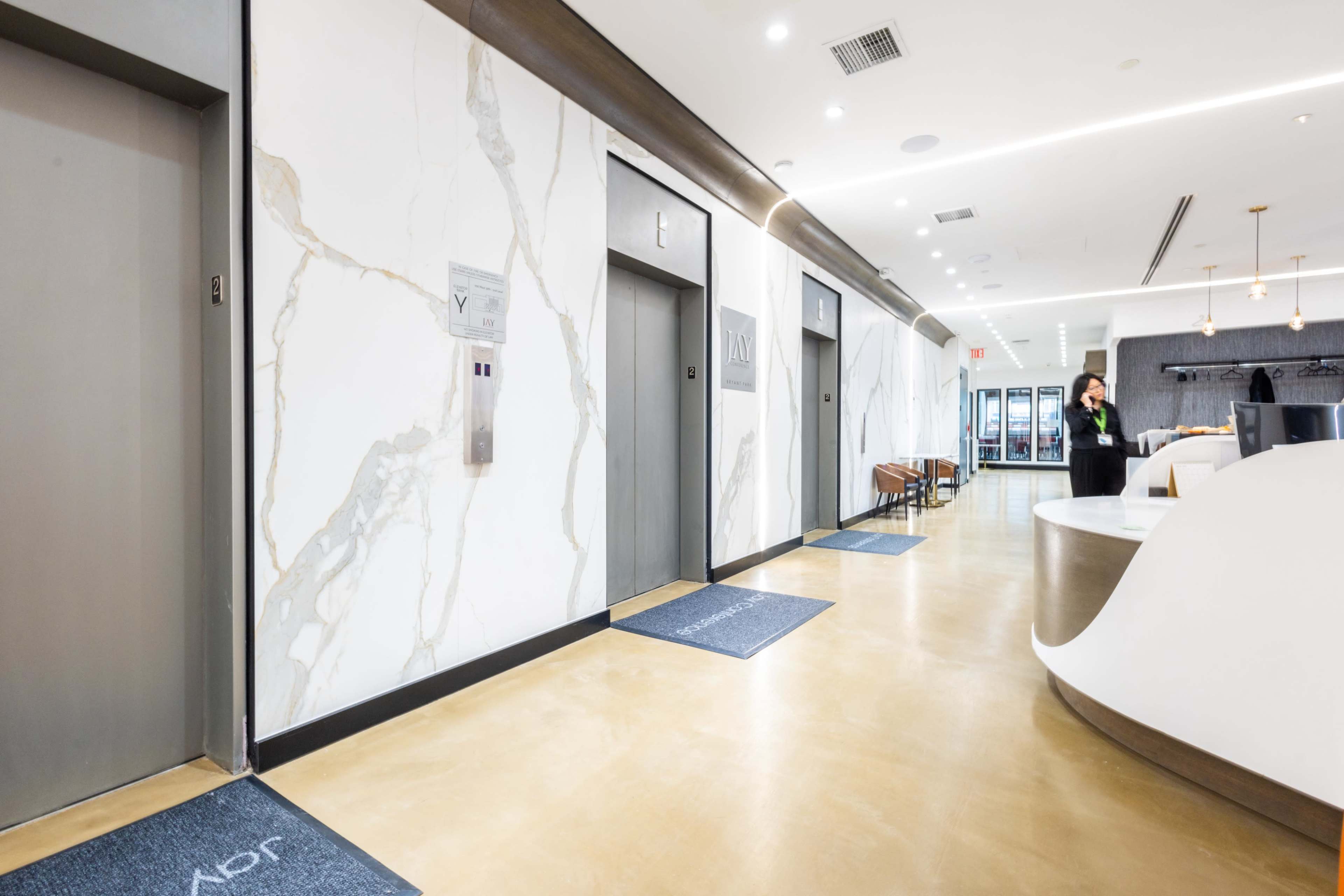 The image shows a modern corridor with elevators on one side, a reception area, and minimalist decor.