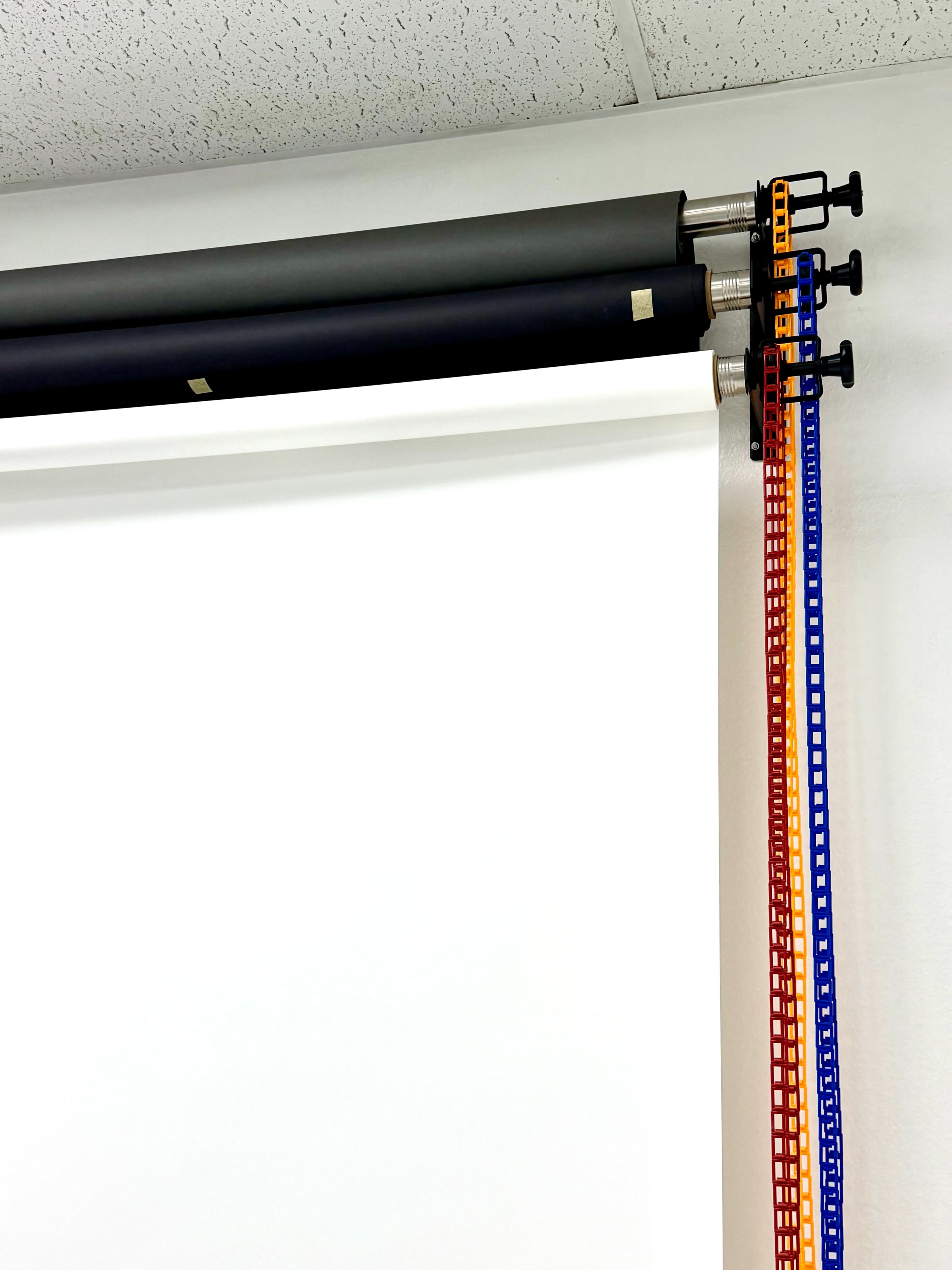 The image shows a wall-mounted roll-up screen with two rolls of black material above a white screen and colored pull cords in orange and blue.