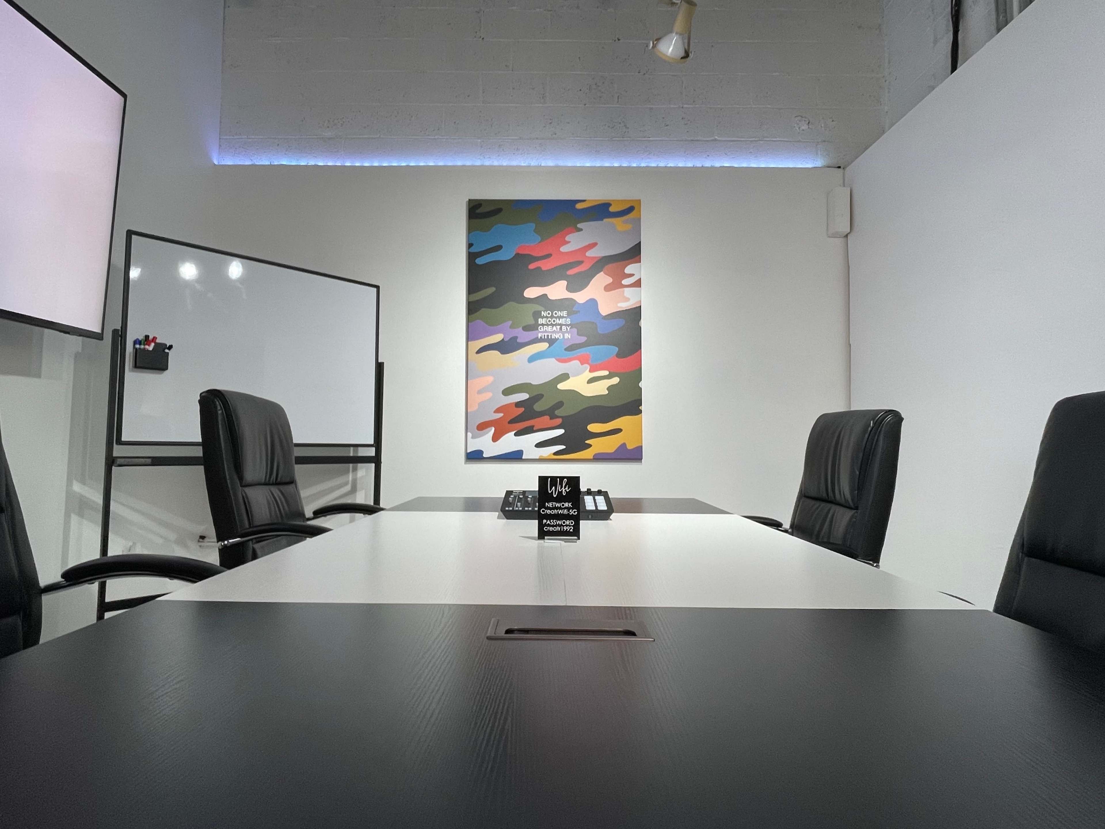 A modern conference room features a long table with black chairs, a wall-mounted screen, and a colorful abstract painting.