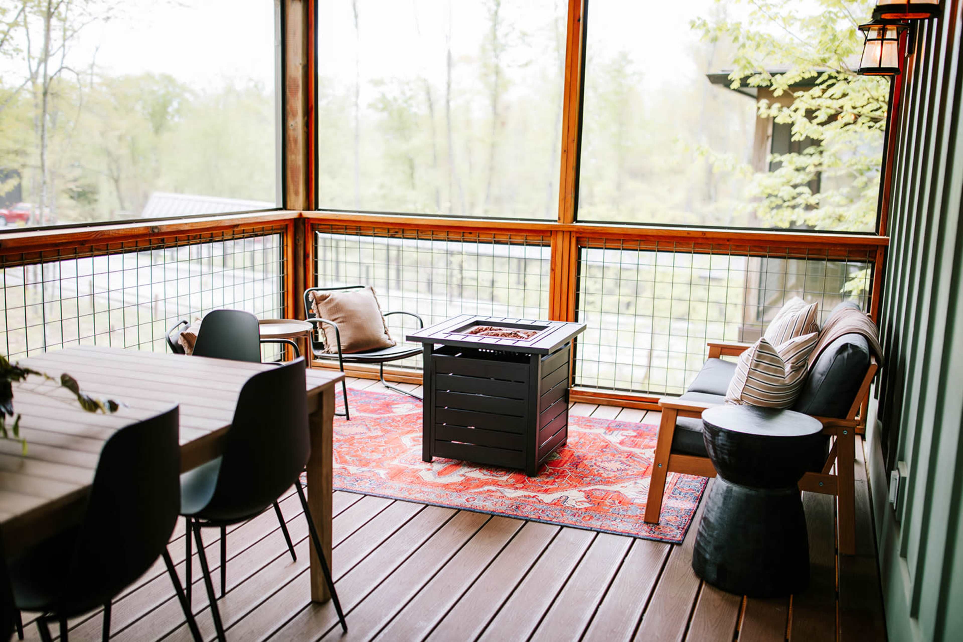 The image shows a screened-in porch with a fire pit table, comfortable seating, and a wooden dining table set against a backdrop of greenery.