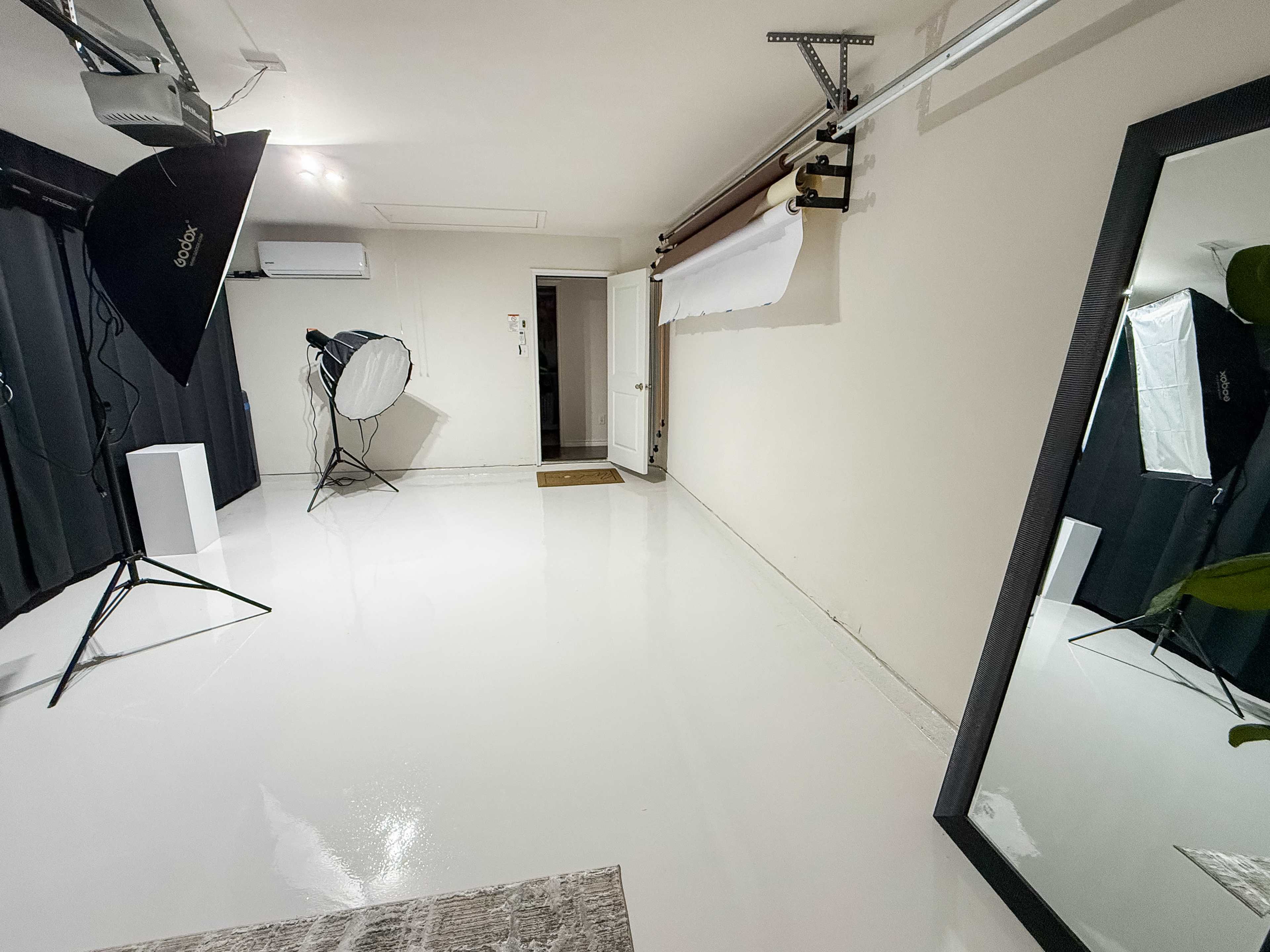 The image shows a photography studio with a shiny white floor, a large mirror, and various lighting equipment set up against plain walls.