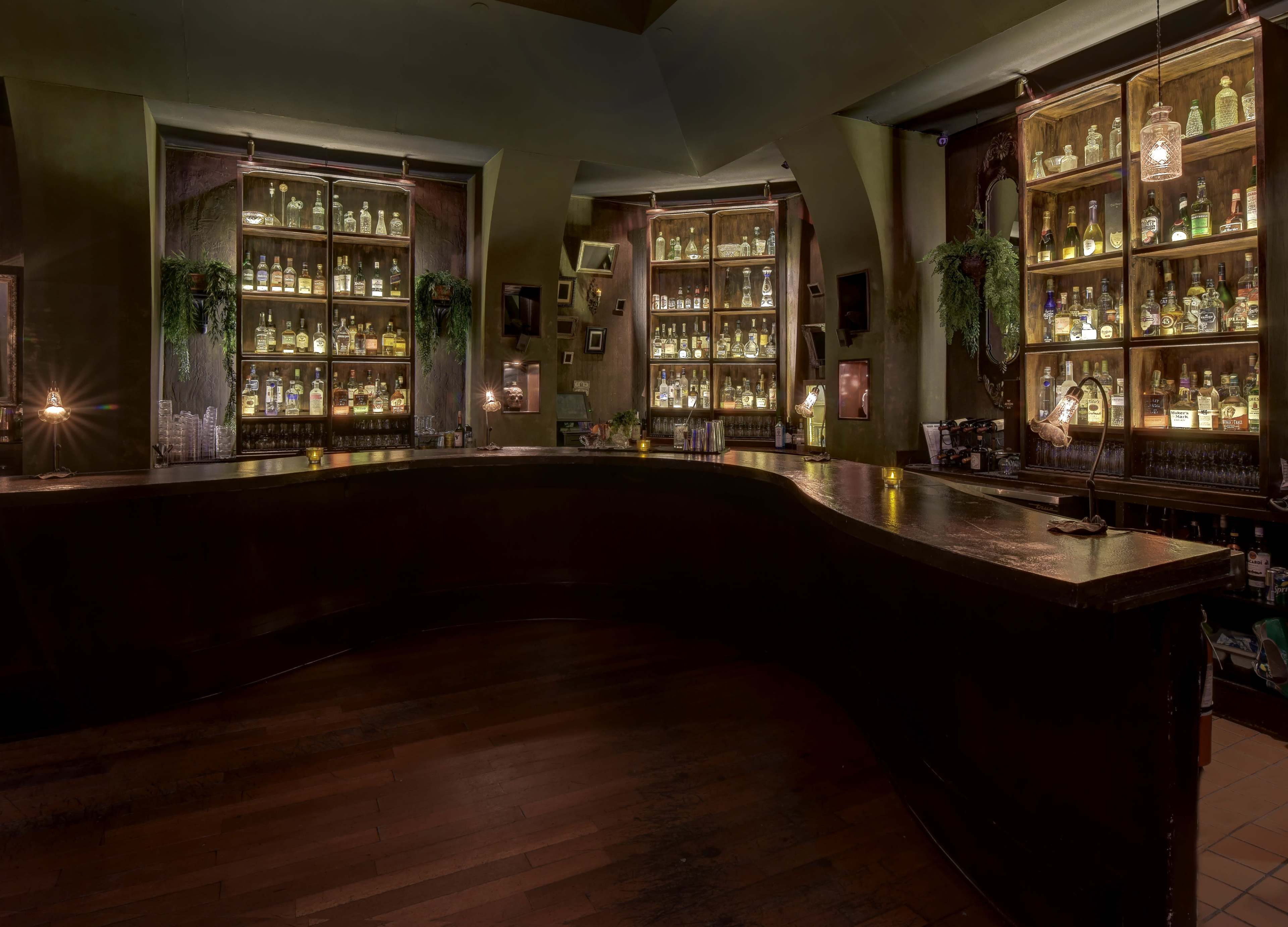 A dimly lit bar with an ornate wooden counter and shelves filled with various bottles of liquor against green walls.