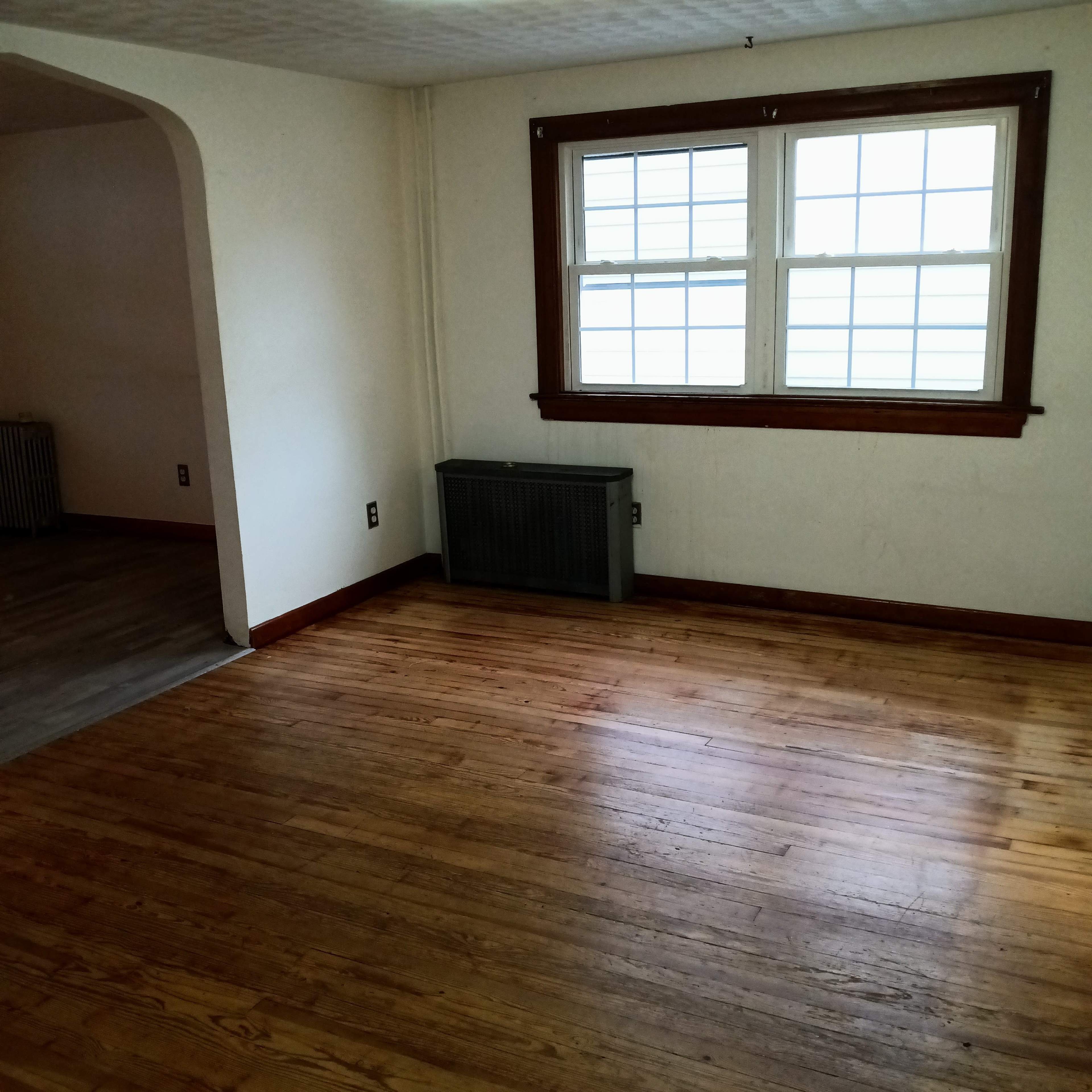 A room with hardwood floors, a large window, and a radiator sits adjacent to another space.