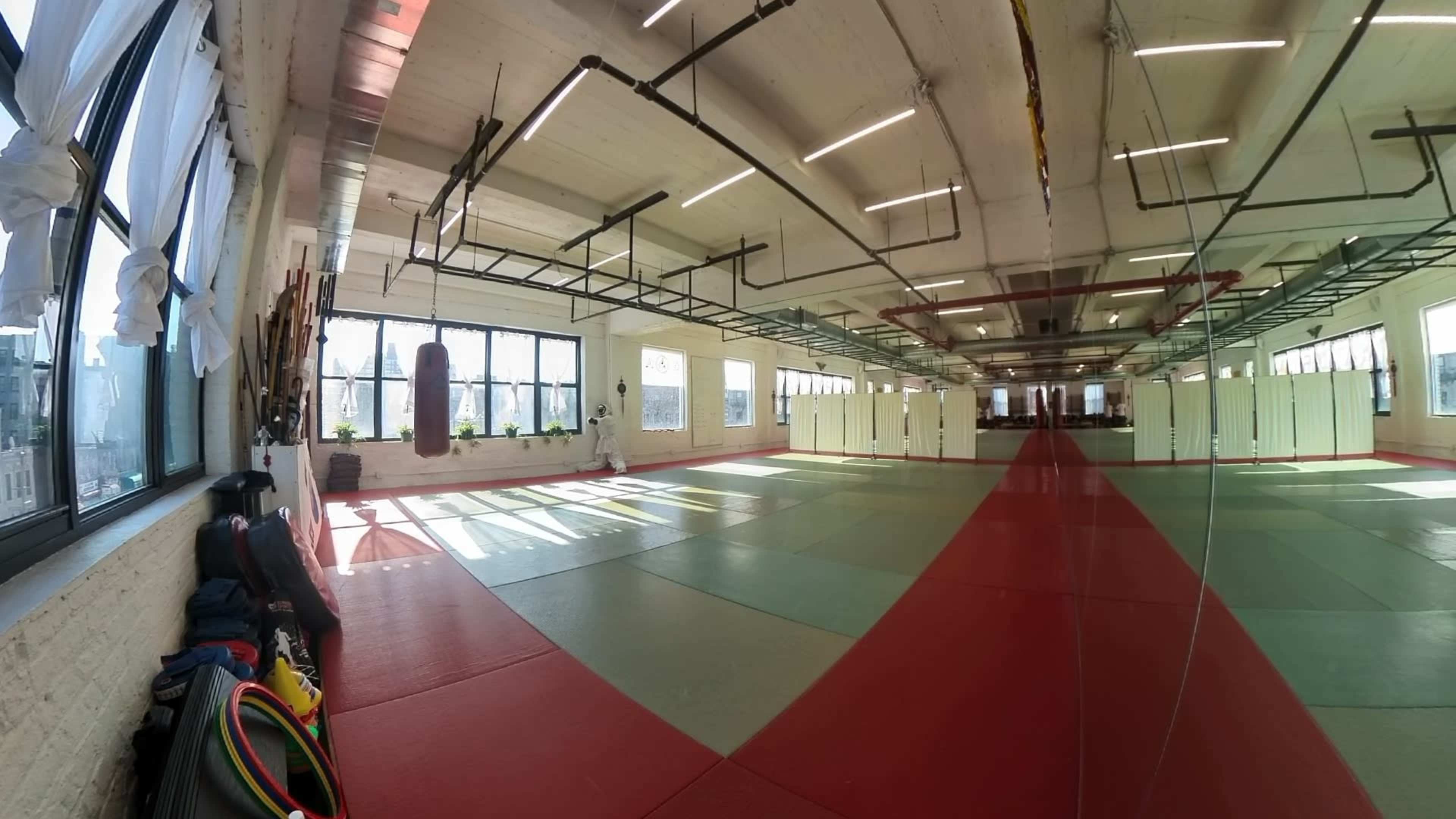 The image shows a spacious training room with red and green mats, large windows letting in natural light, and a mirrored wall reflecting the area.