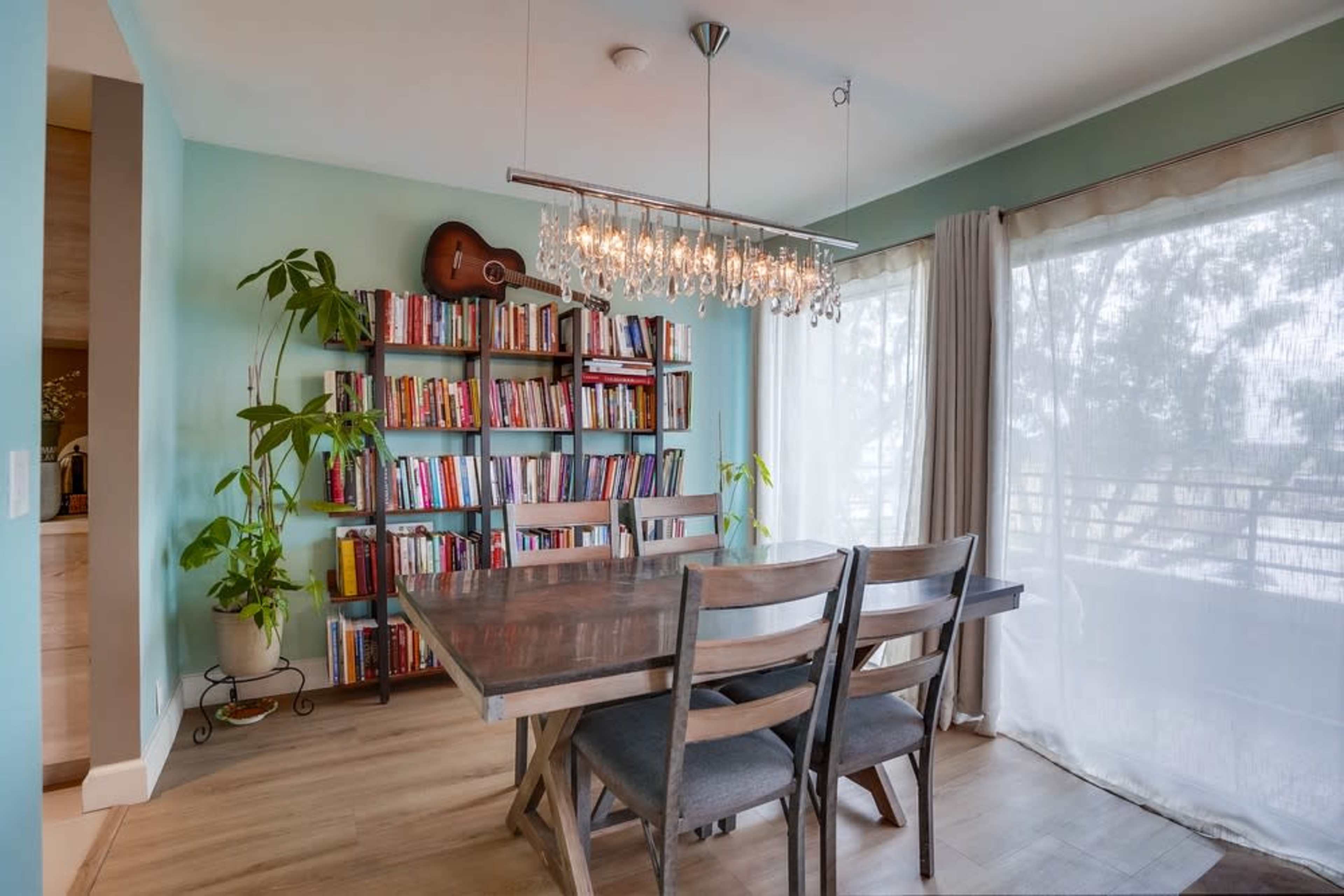 A dining area features a wooden table with four chairs, surrounded by a bookshelf filled with books, a guitar hanging on the wall, and large windows allowing natural light to enter.