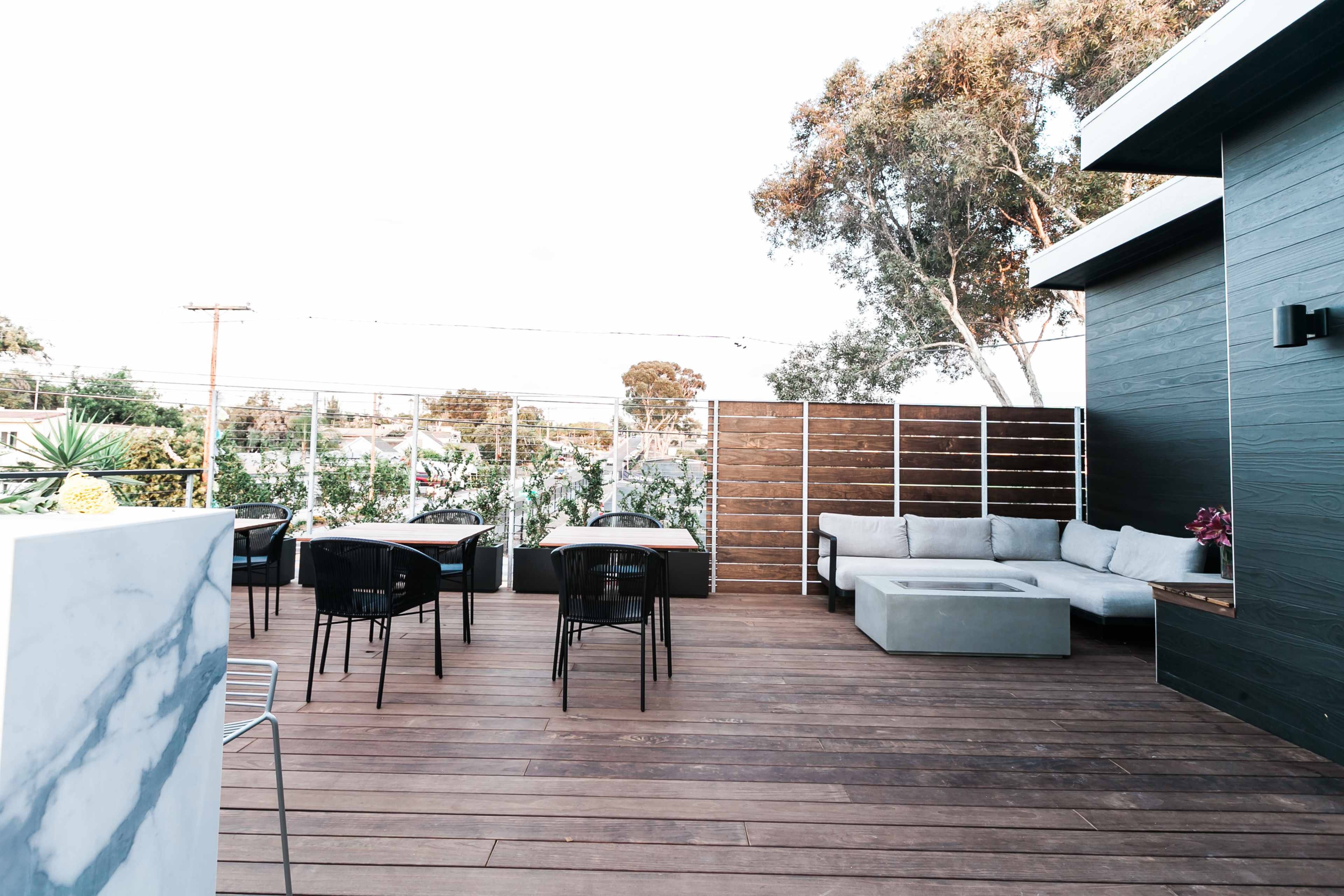 The image shows a wooden deck area furnished with tables and seating surrounded by greenery and modern fencing.