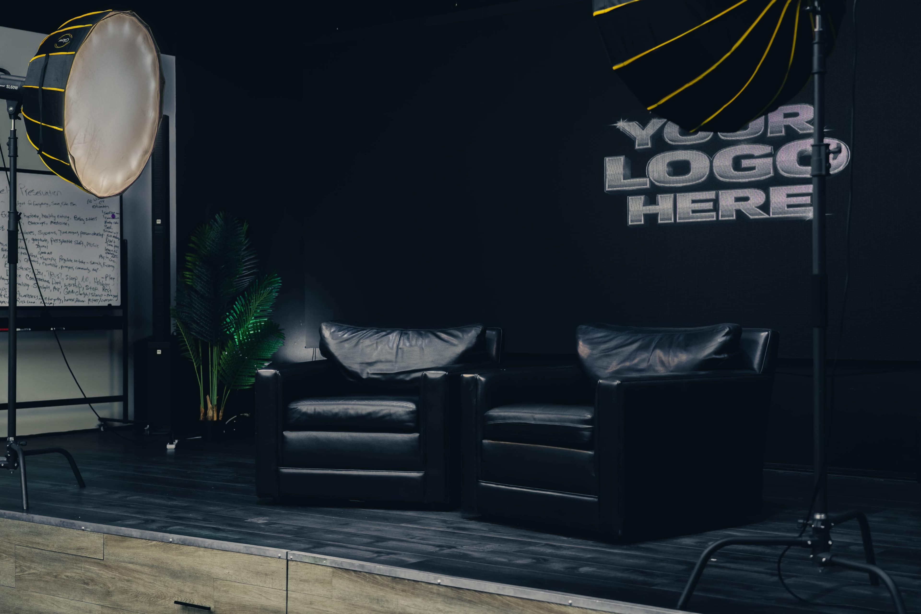 The image shows a minimalist studio setup with two black leather chairs, a large light source, and a backdrop featuring a placeholder for a logo.