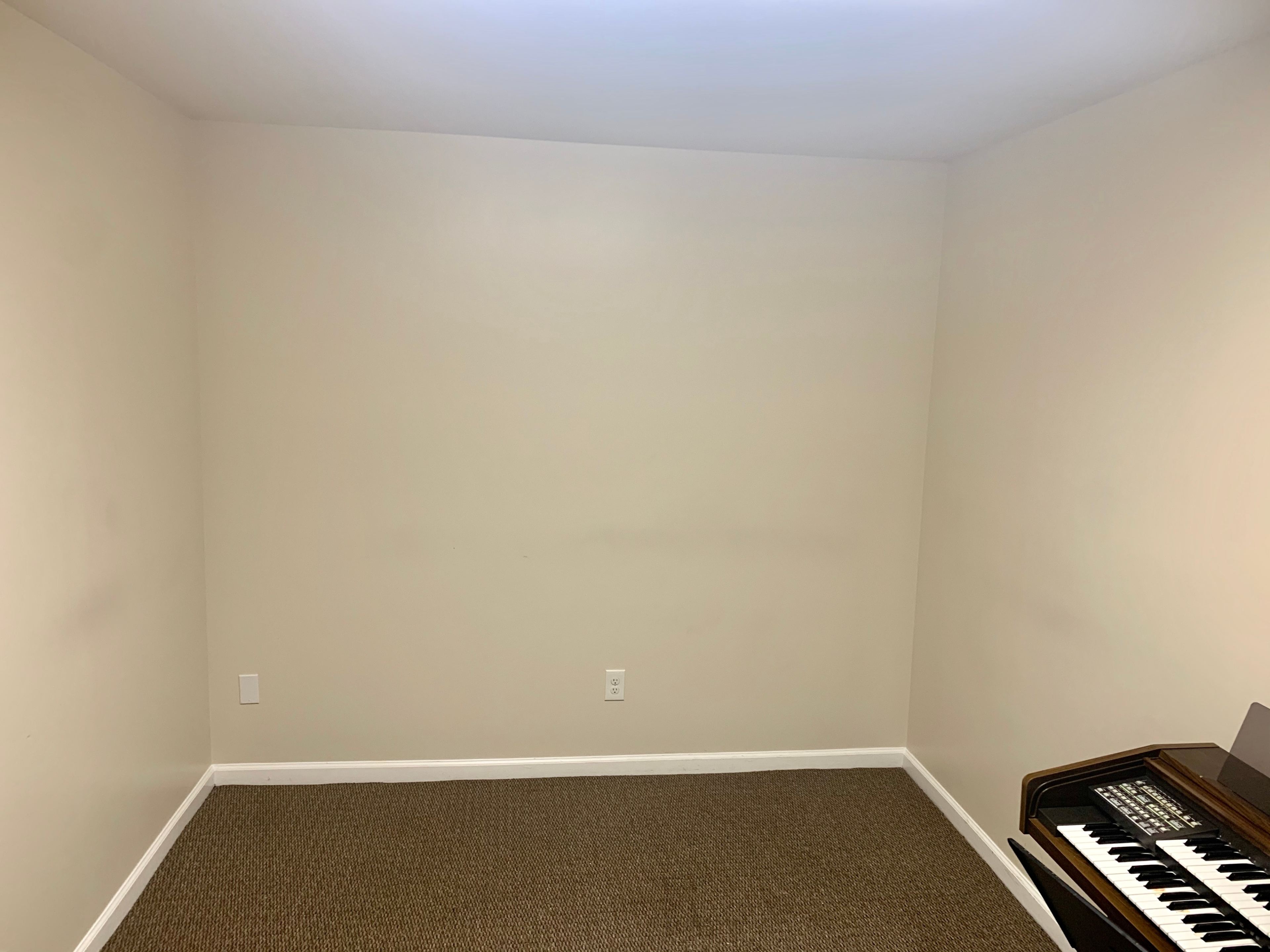 An empty room with beige walls and a carpeted floor, featuring a keyboard on the right side.