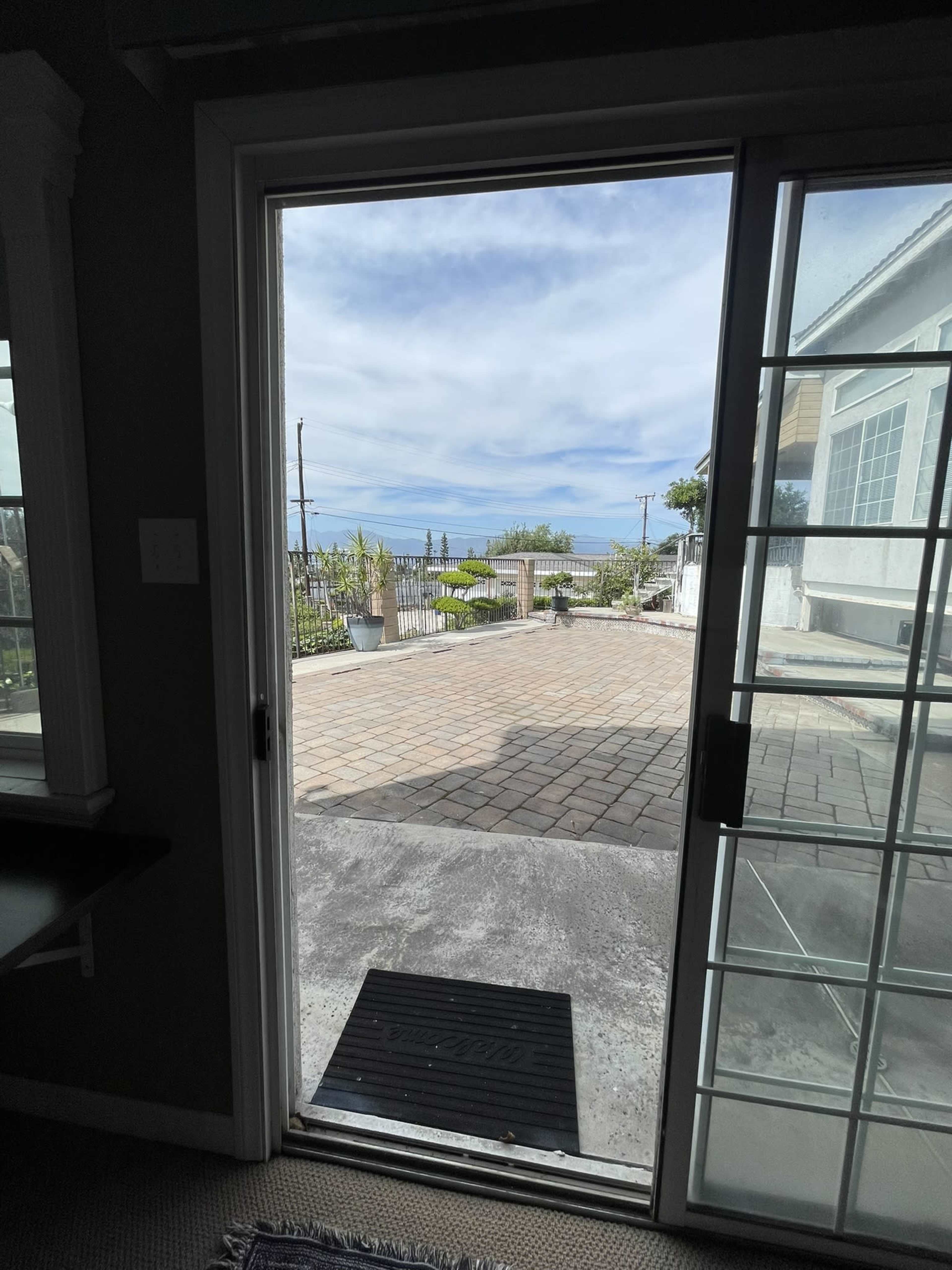 The image shows an open door leading to a paved outdoor area with a view of trees and a cloudy sky.