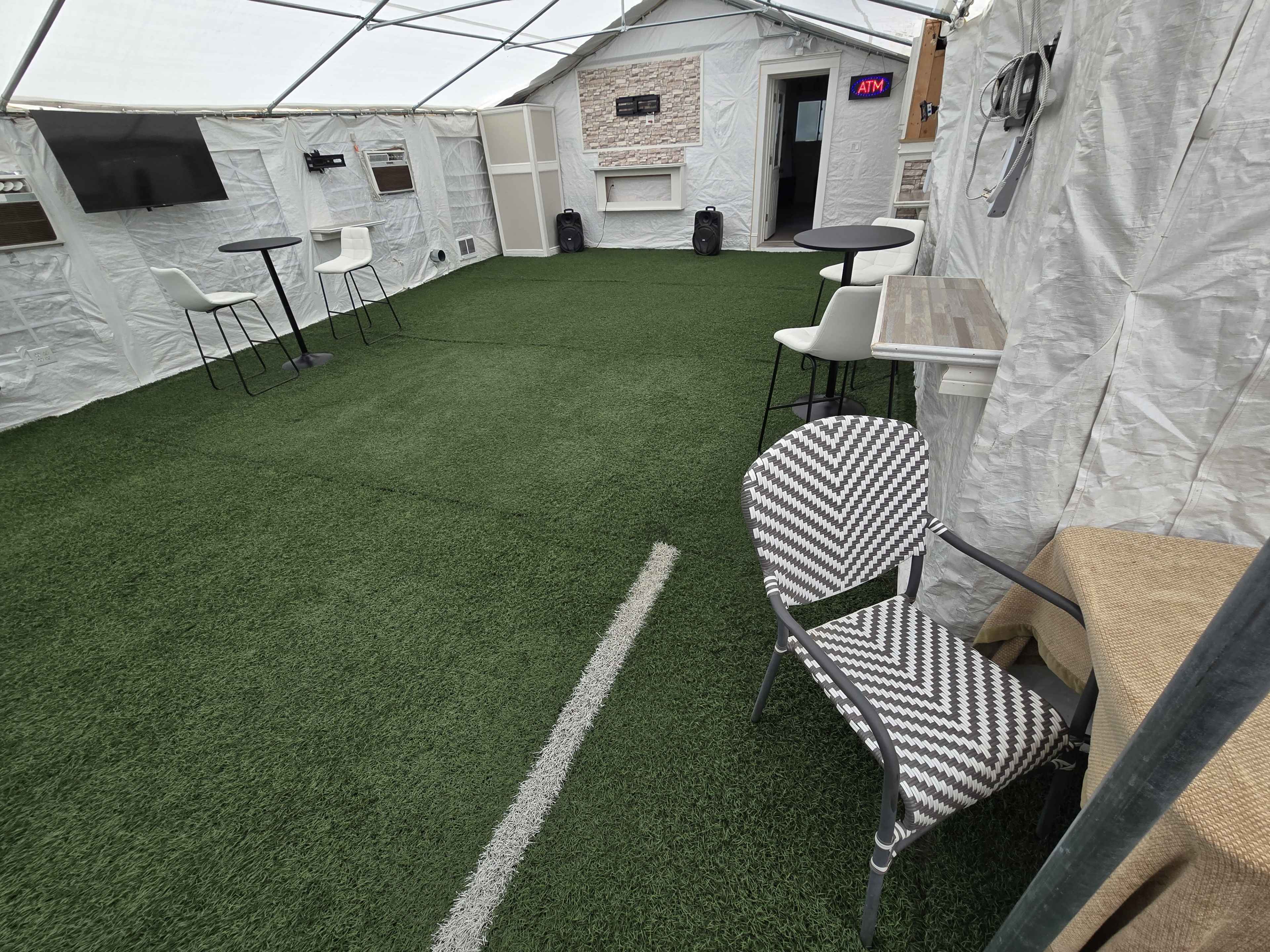 The image shows an indoor space with artificial turf flooring, featuring tables, chairs, and a television mounted on the wall.