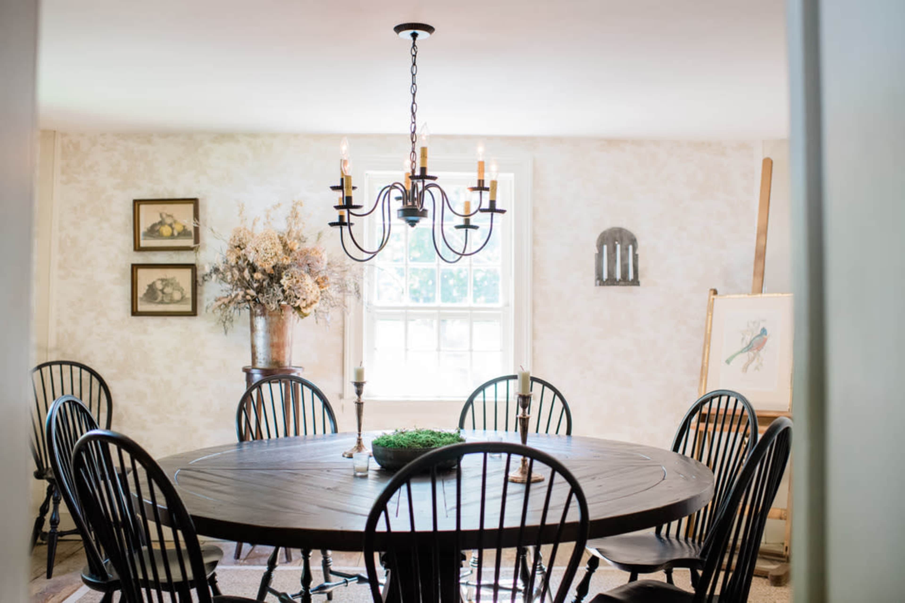 A round wooden dining table is surrounded by black spindle-back chairs in a softly lit room featuring floral decor and artwork.