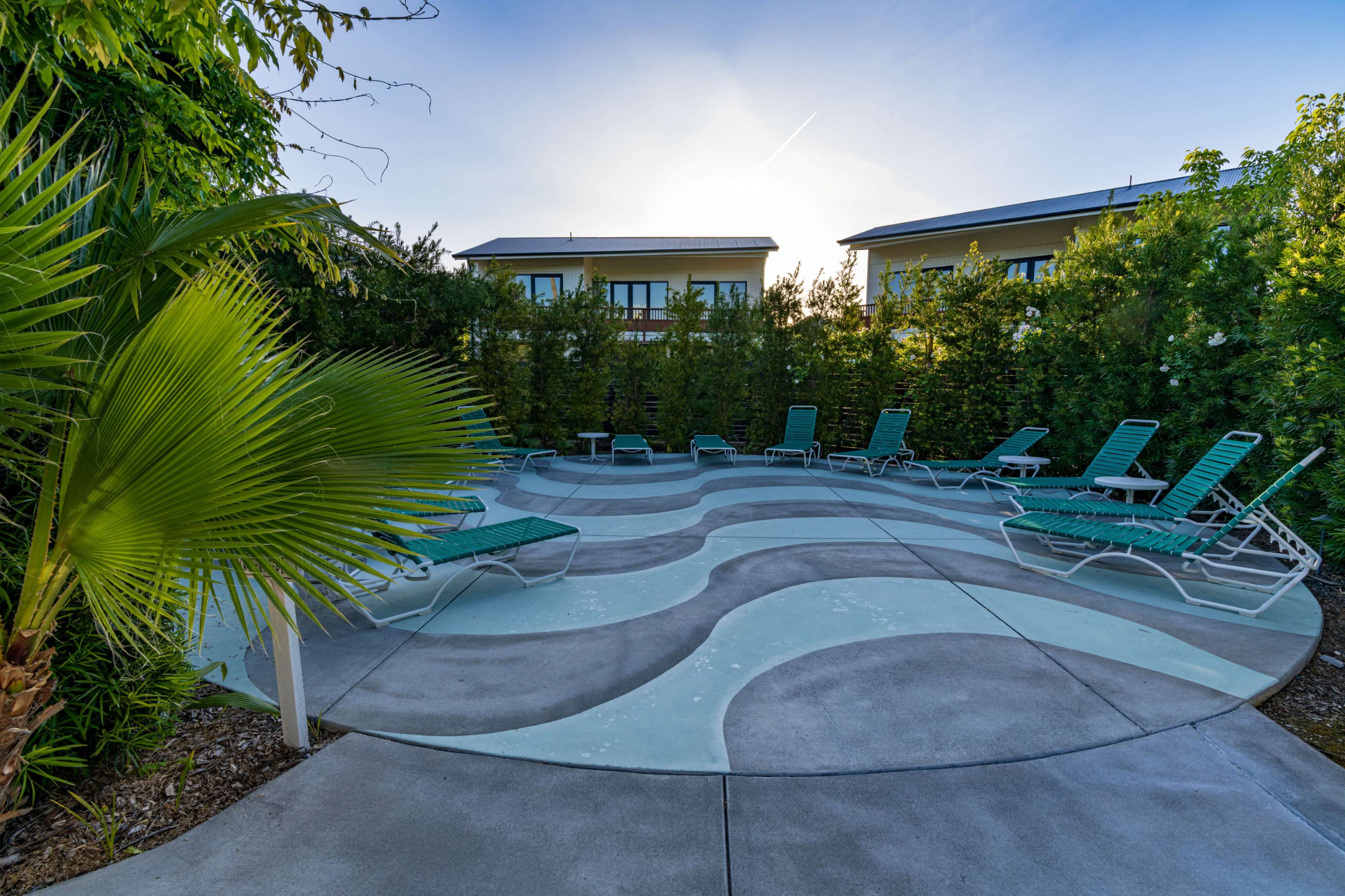 The image shows a pool area with curved, wave-patterned concrete and several green lounge chairs surrounded by lush greenery.