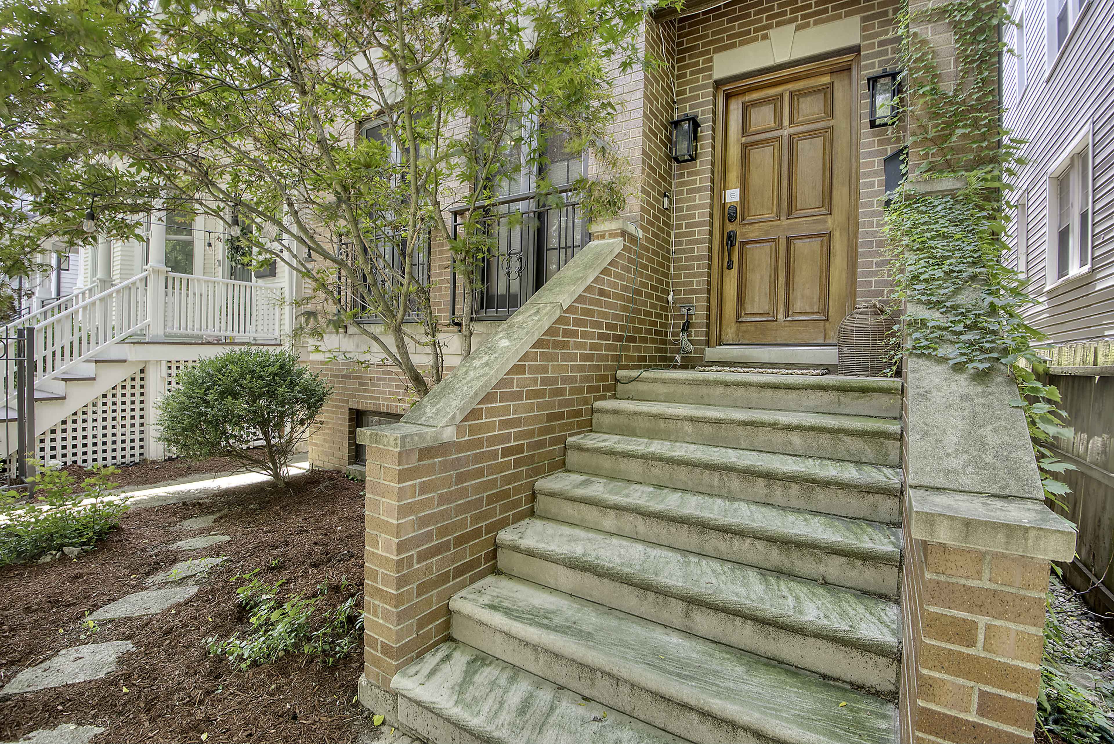 The image shows a brick stairway leading to a wooden front door, surrounded by green foliage and a landscaped pathway.
