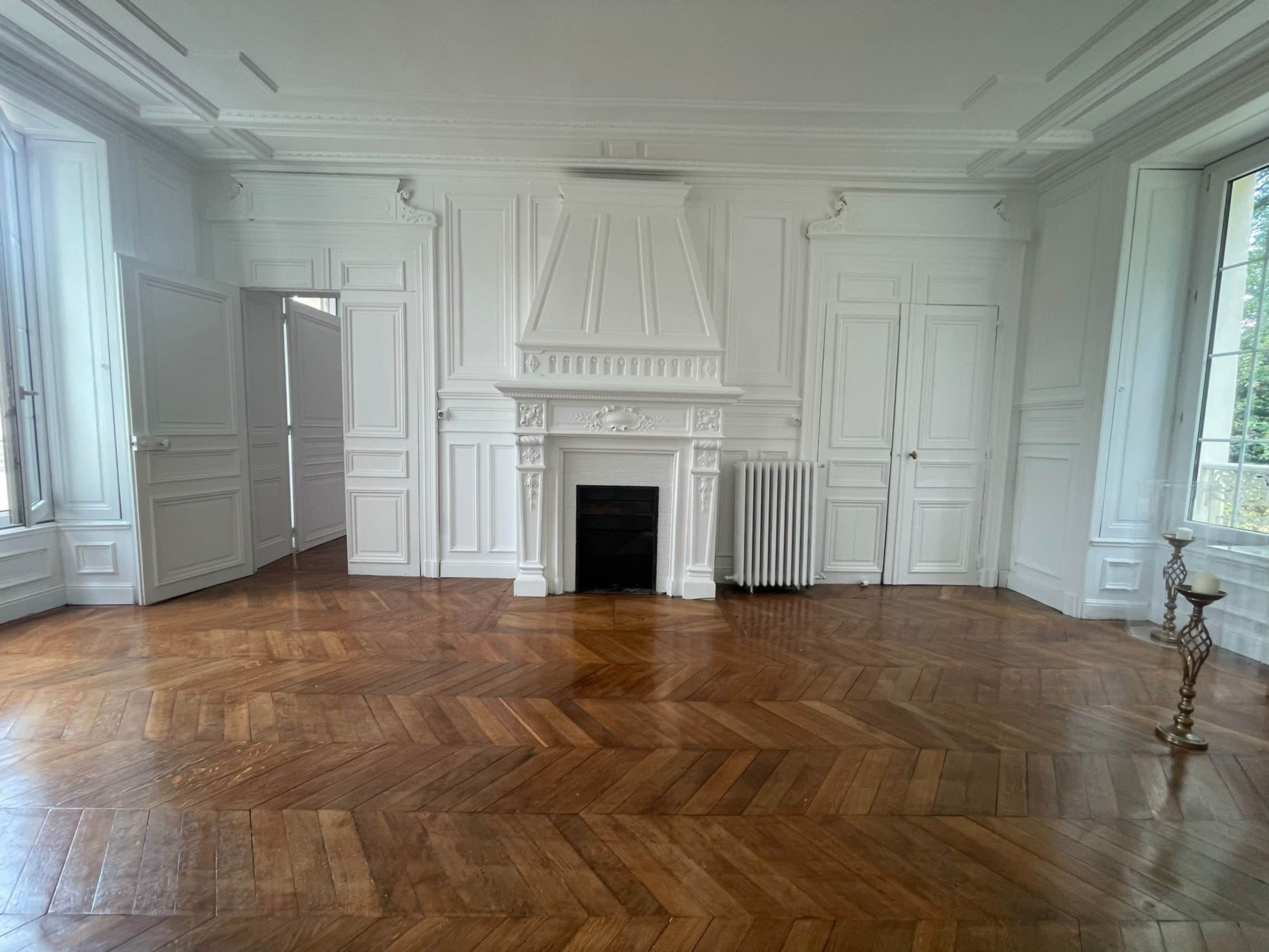 A spacious room with white-paneled walls, a decorative fireplace, and a herringbone-patterned wooden floor.