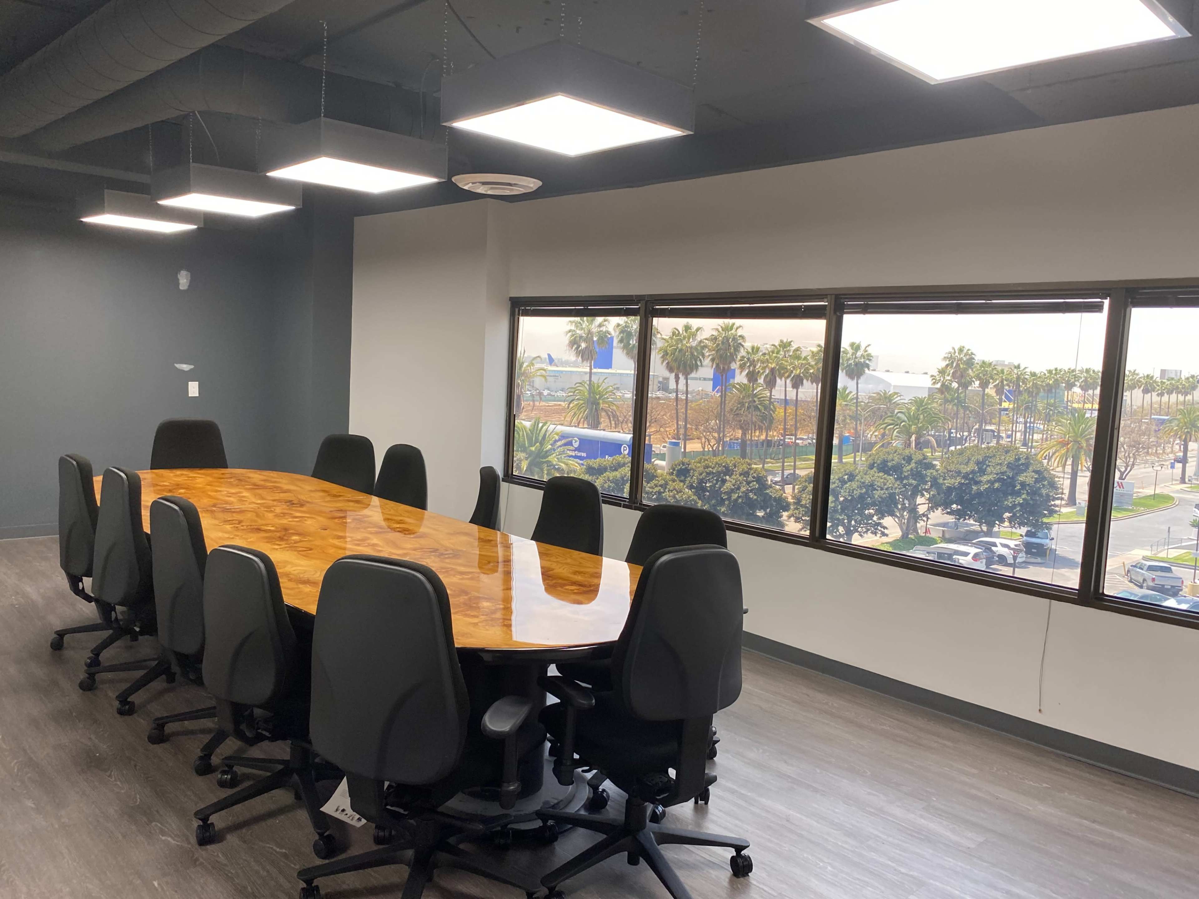 A conference room features a large wooden table surrounded by black office chairs, with large windows showing an exterior view of palm trees and a parking area.