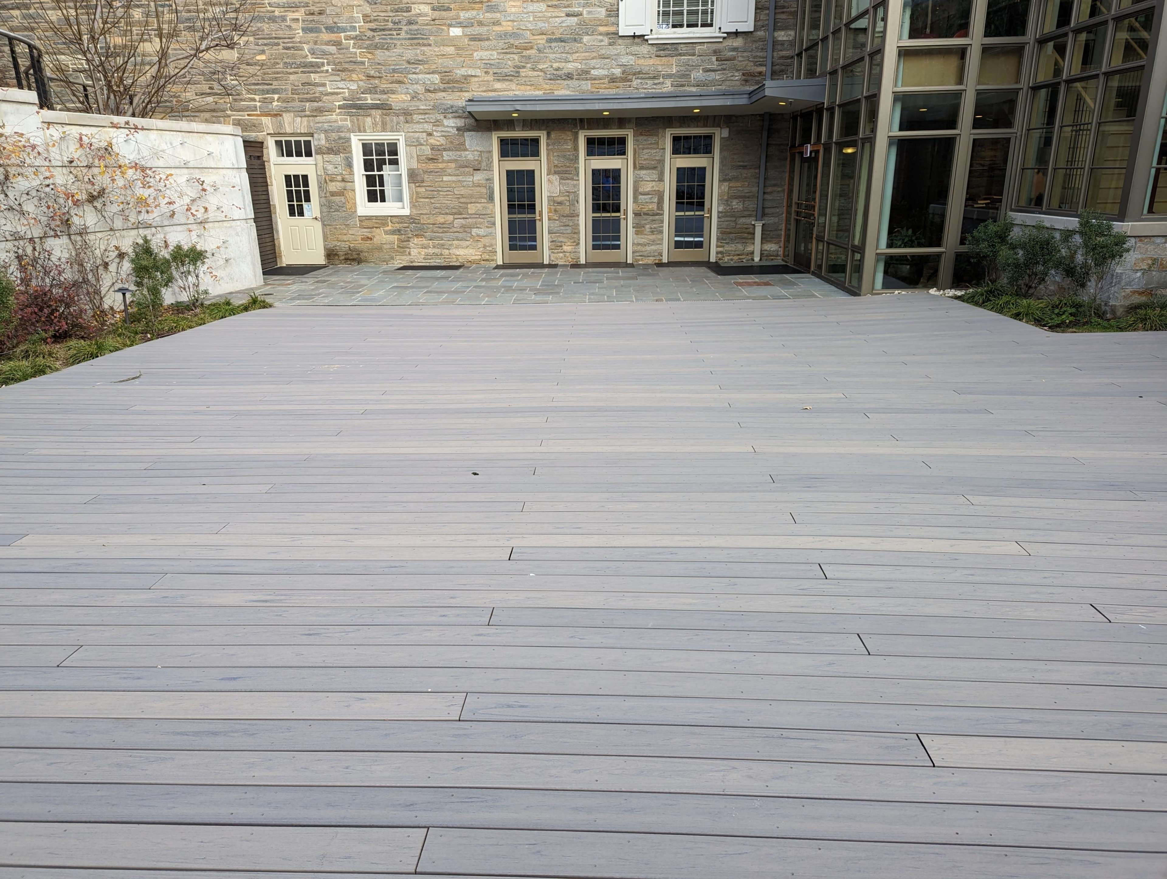 A wooden deck area with gray planks, leading to a stone building with large windows and a set of doors.
