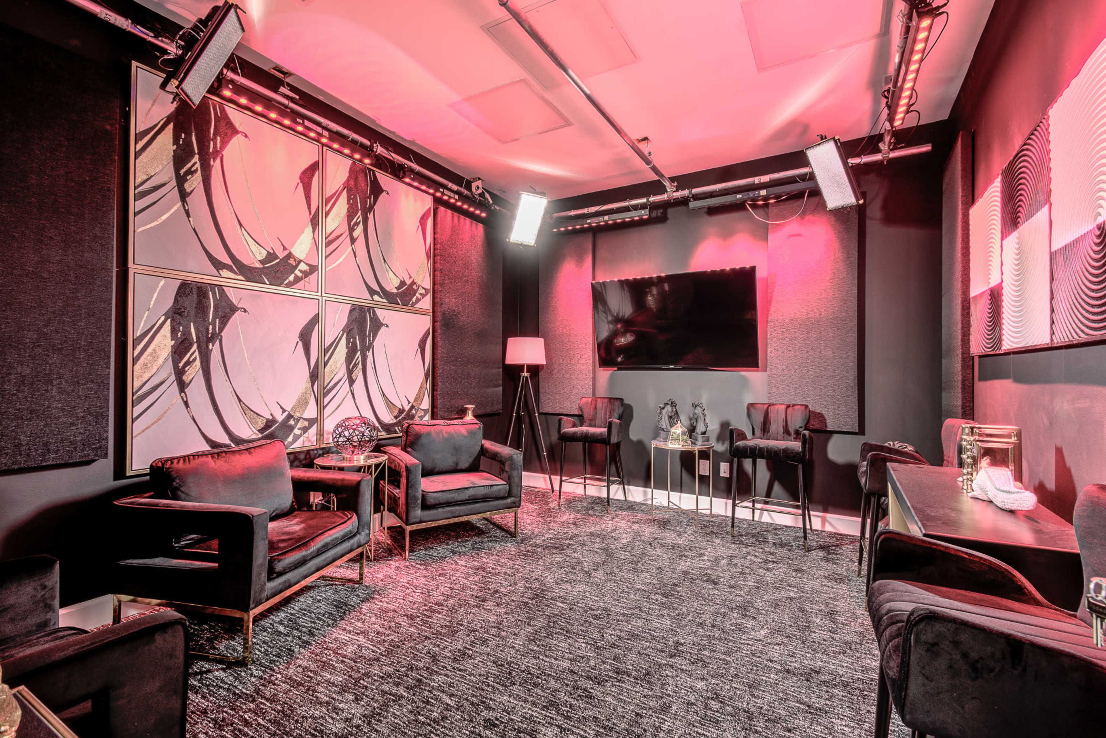 The image shows a modern lounge area with black furniture, a large television on the wall, and artistic wall decor illuminated with soft pink lighting.
