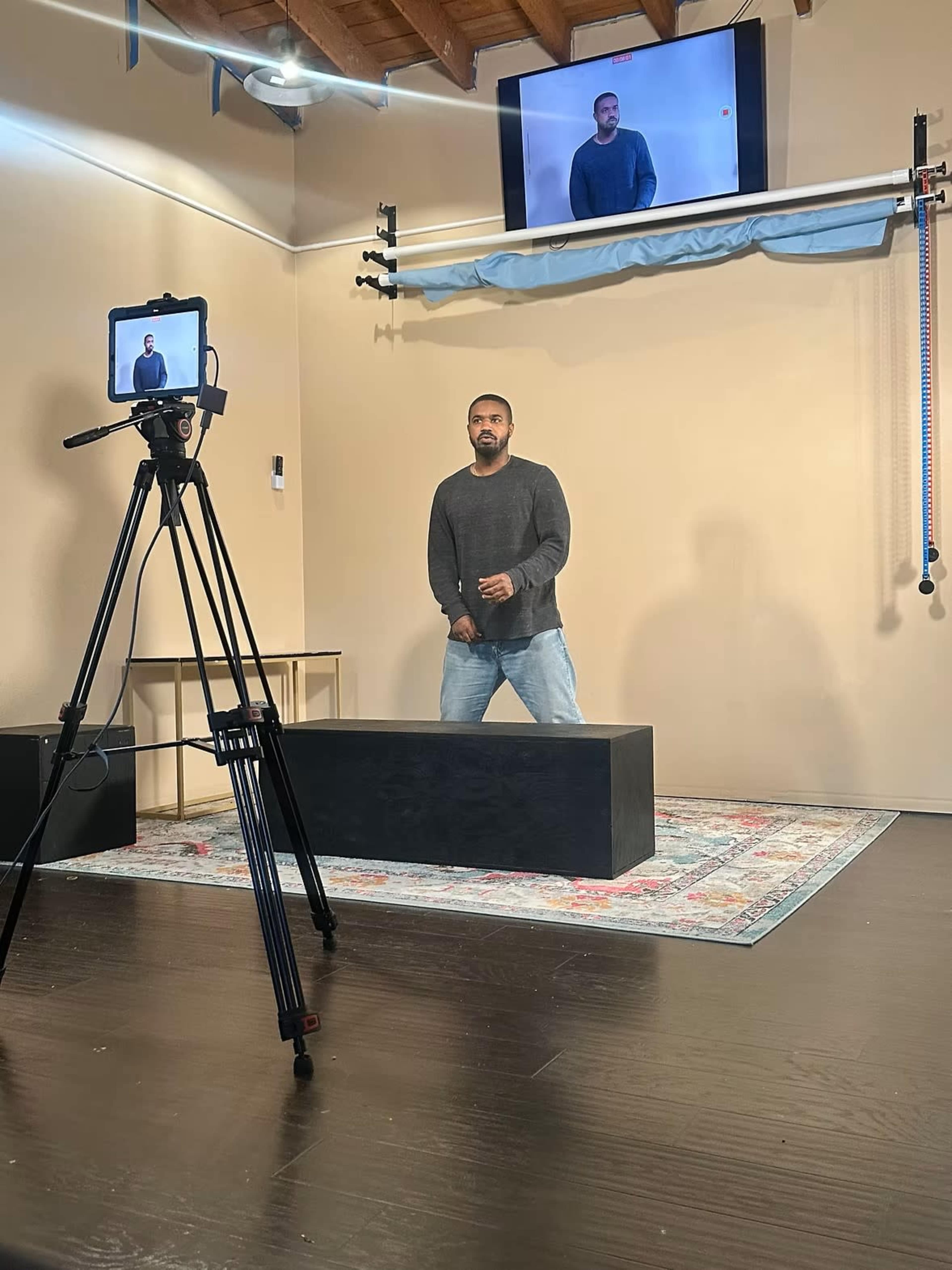 A person stands on a black platform in front of a camera, with a monitor displaying their image in a well-lit room.