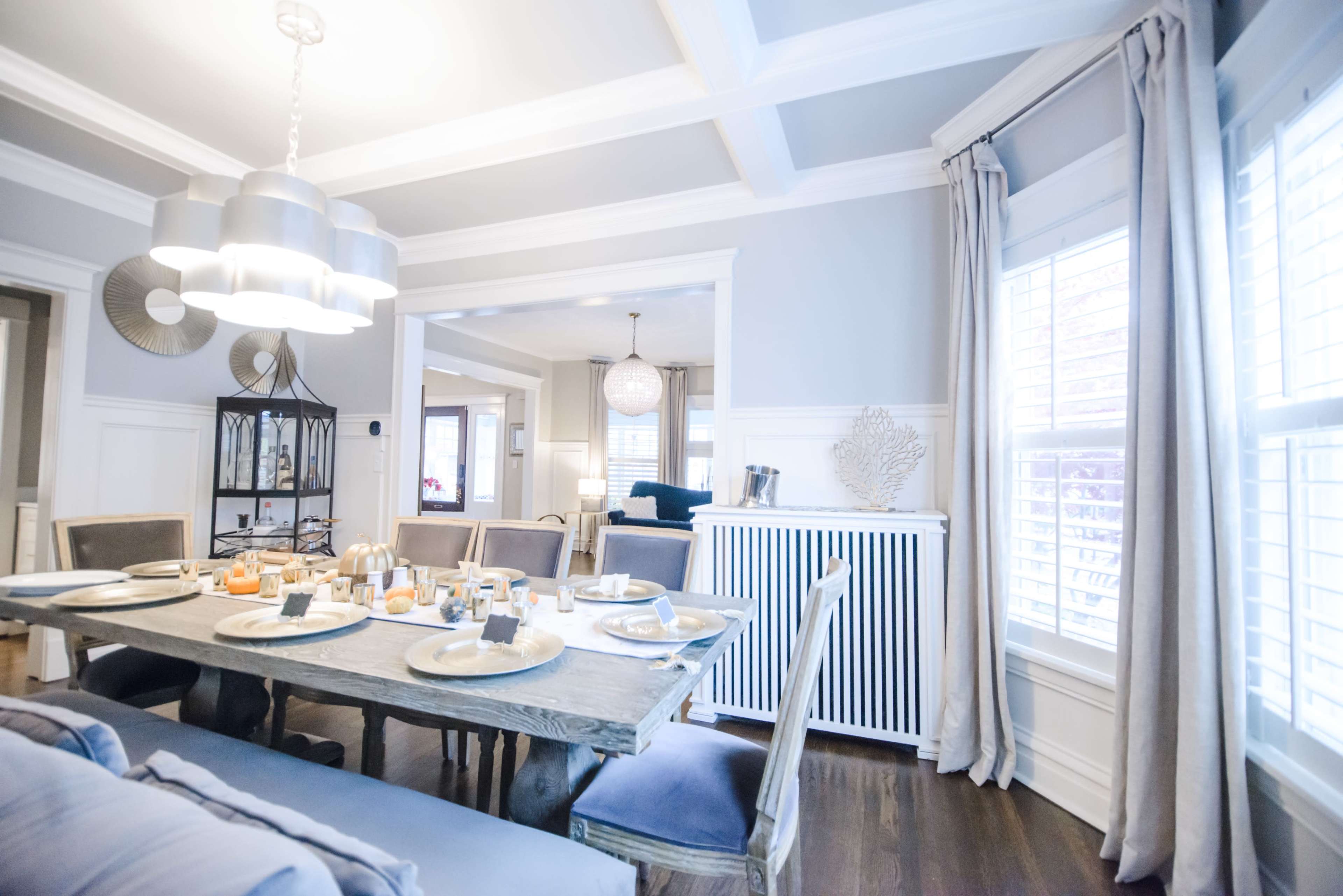 A dining room features a wooden table set with plates and decorative items, surrounded by cushioned chairs, with windows allowing natural light and a view into an adjoining living area.