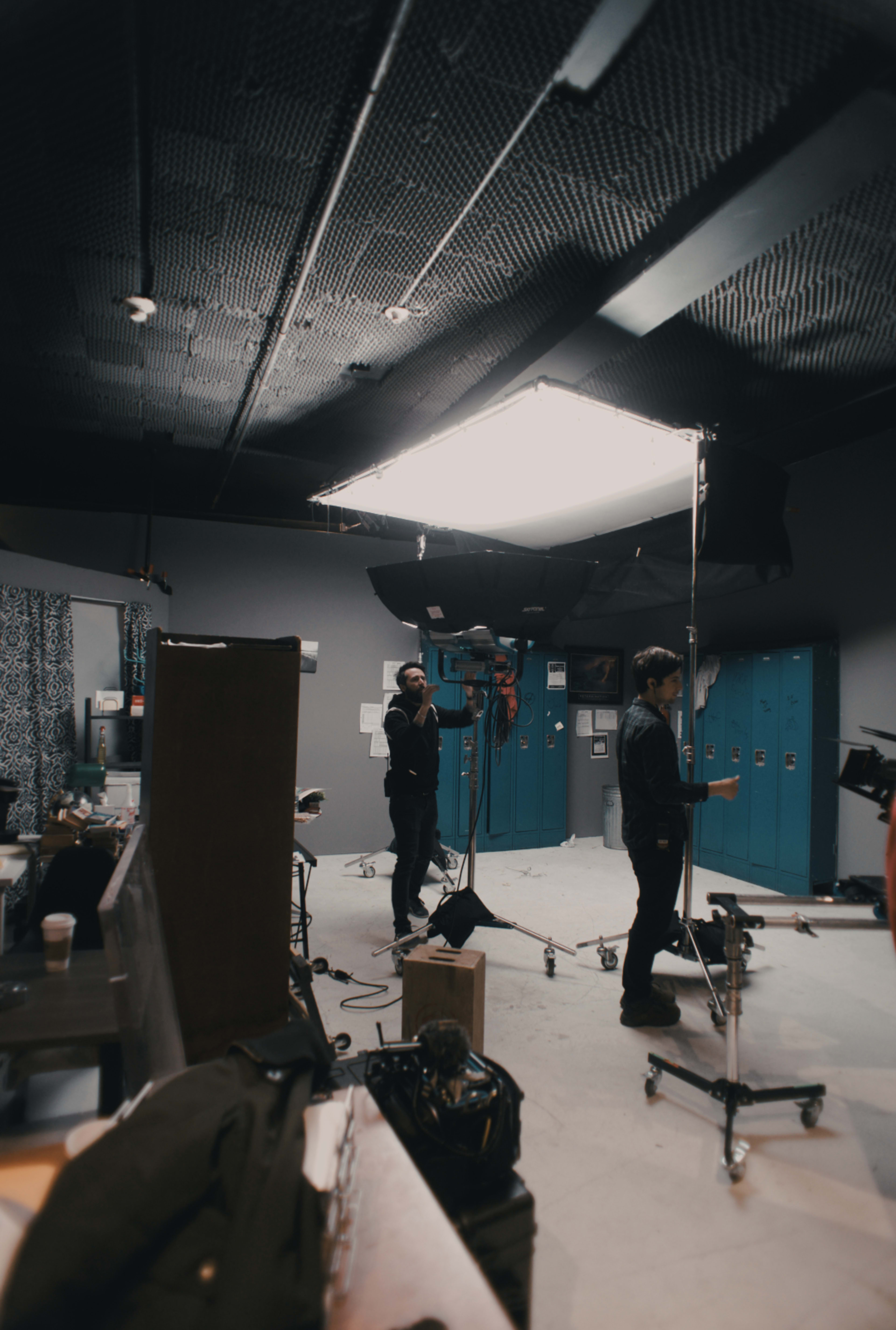 A film crew operates in a studio with blue lockers and various equipment set up for shooting.