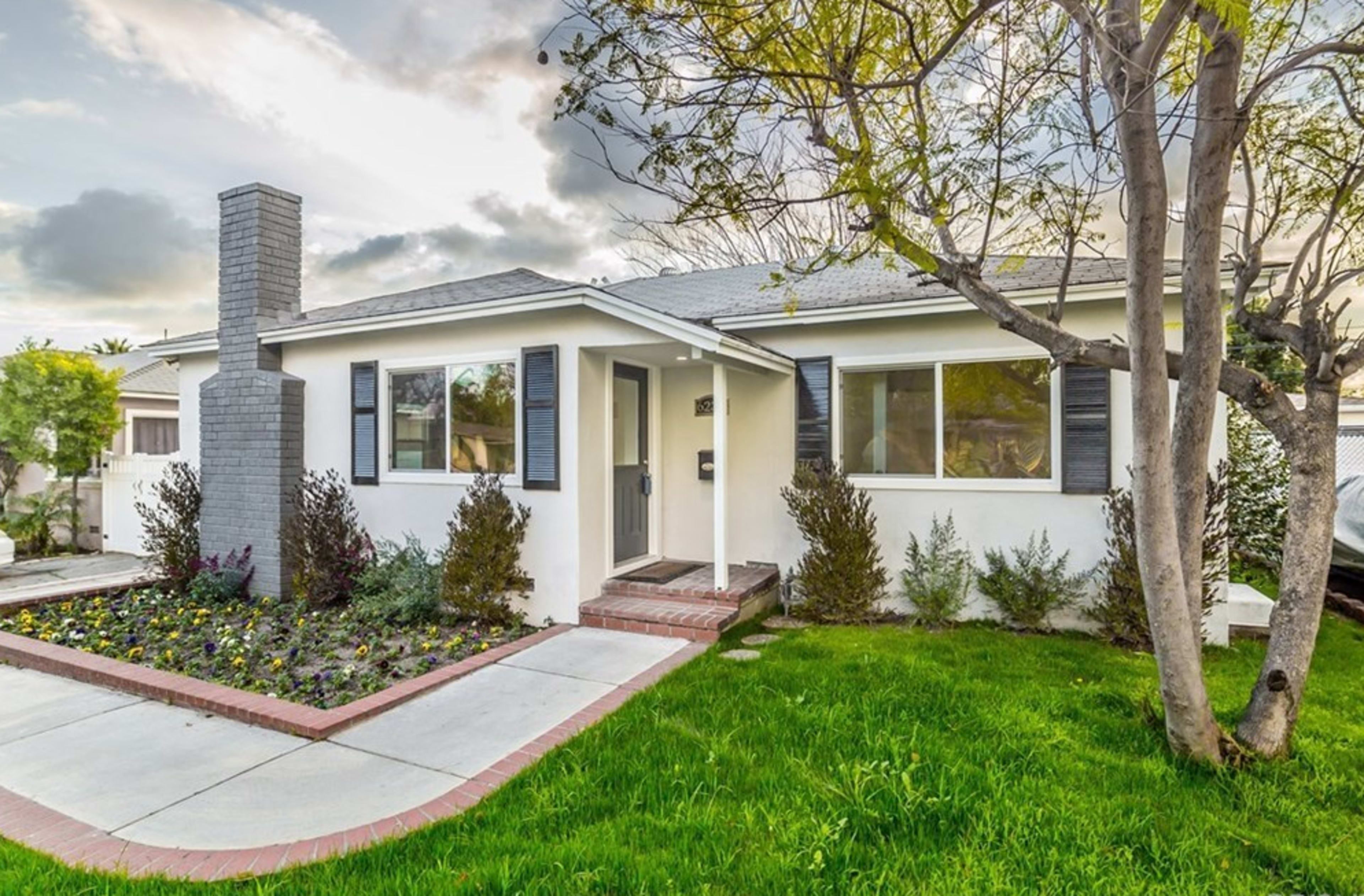 A single-story house with a gray chimney, black shutters, and a well-maintained front yard featuring a flower bed and a sloped green lawn.
