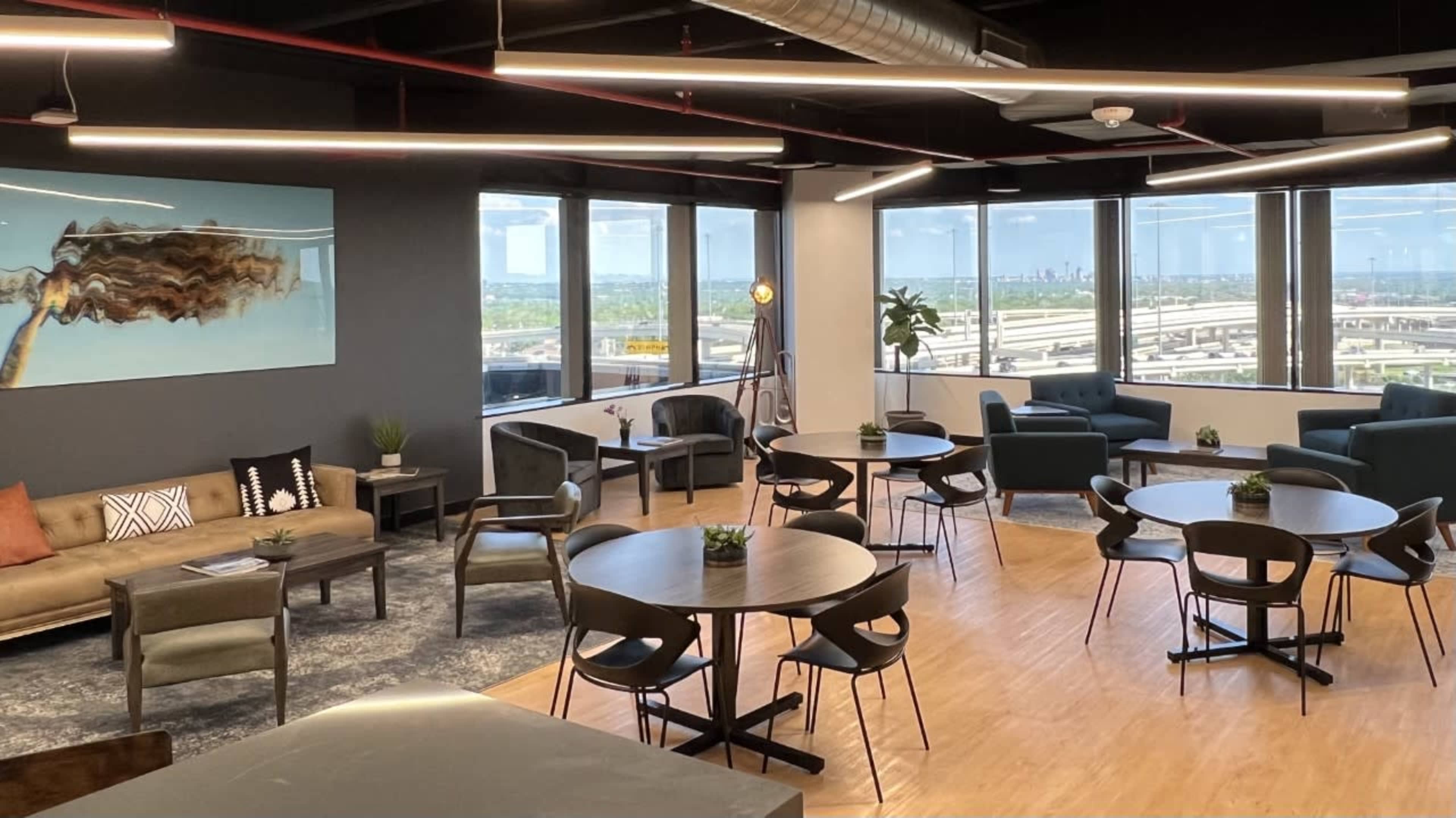 The image shows a modern office lounge with seating areas, round tables, and large windows offering a view of the cityscape.