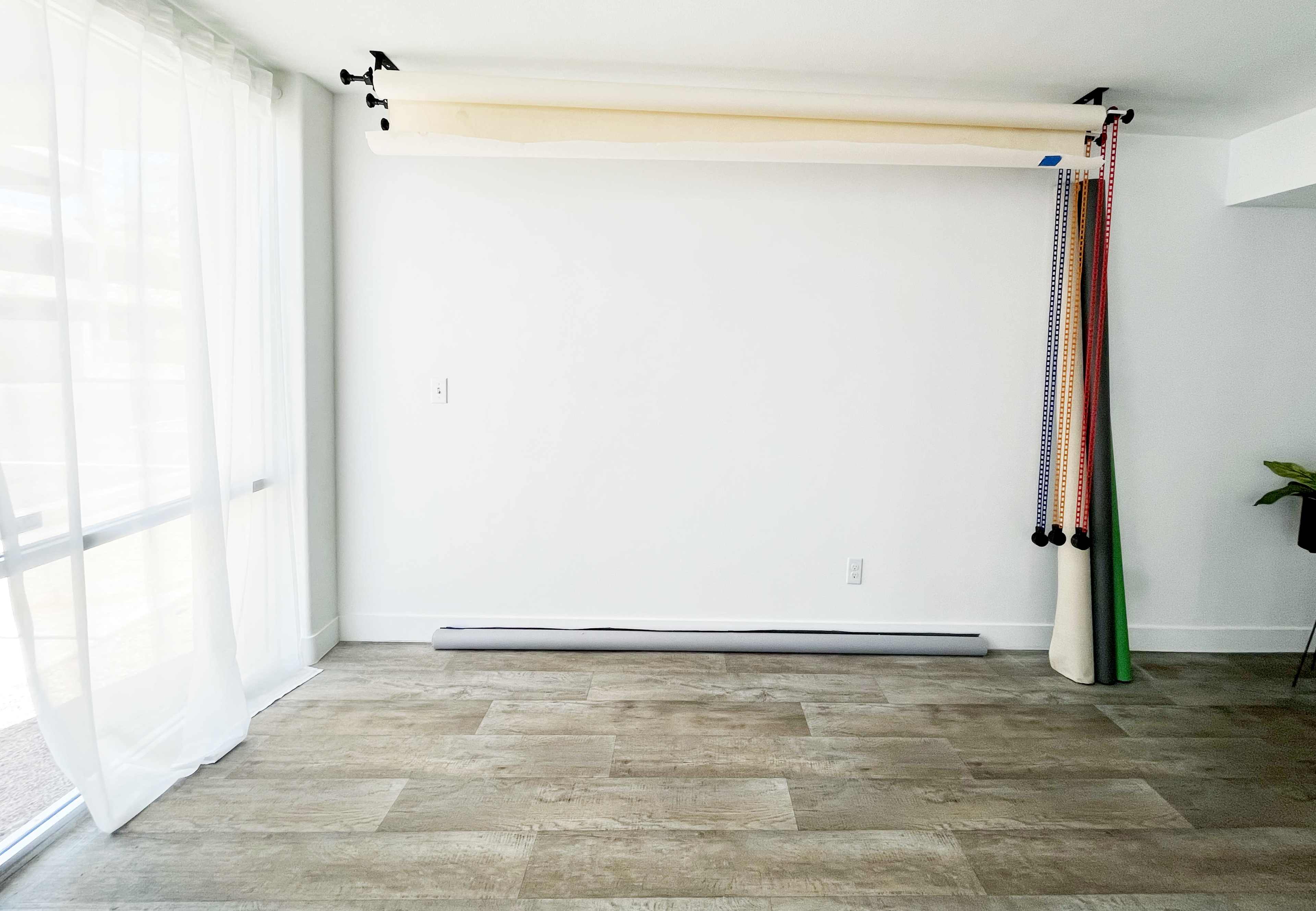 A room with a white wall, a roller blind hanging above exercise bands, and a window with sheer white curtains.